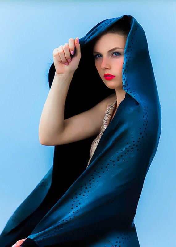 Woman with red lips wrapped in teal blue satin hooded fabric against light blue background.