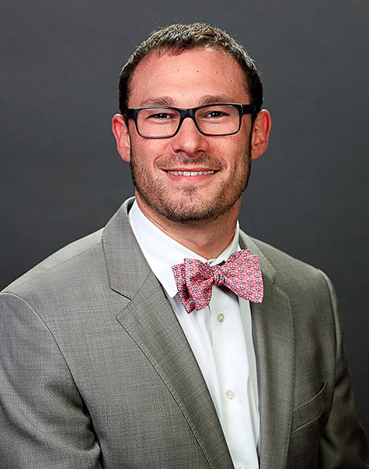 Professional man in gray suit and pink bow tie with black glasses smiling for a business portrait photo.