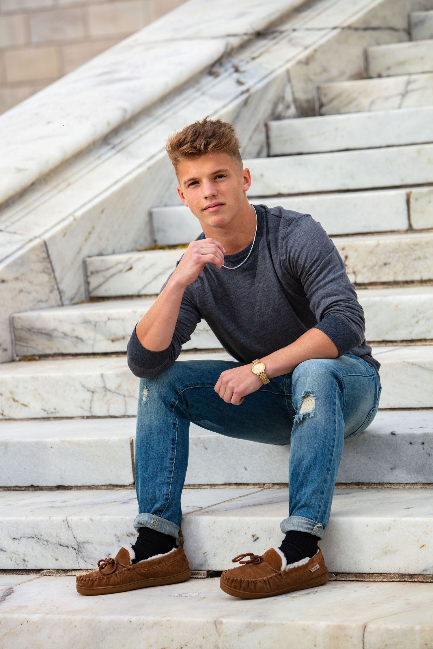 Teen boy in gray sweater and jeans sitting on marble steps in casual senior portrait photo.