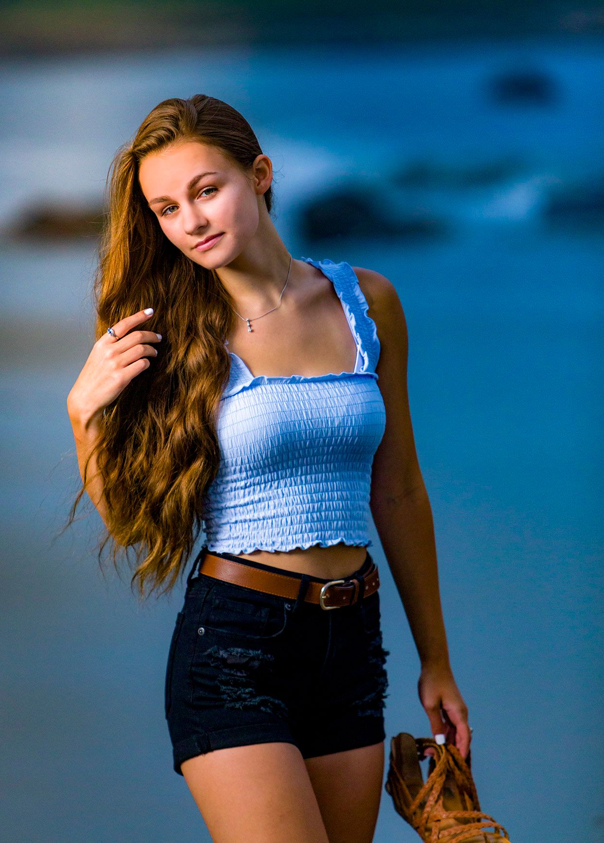 Teen girl with long wavy hair in blue crop top and denim shorts posing at ocean beach.