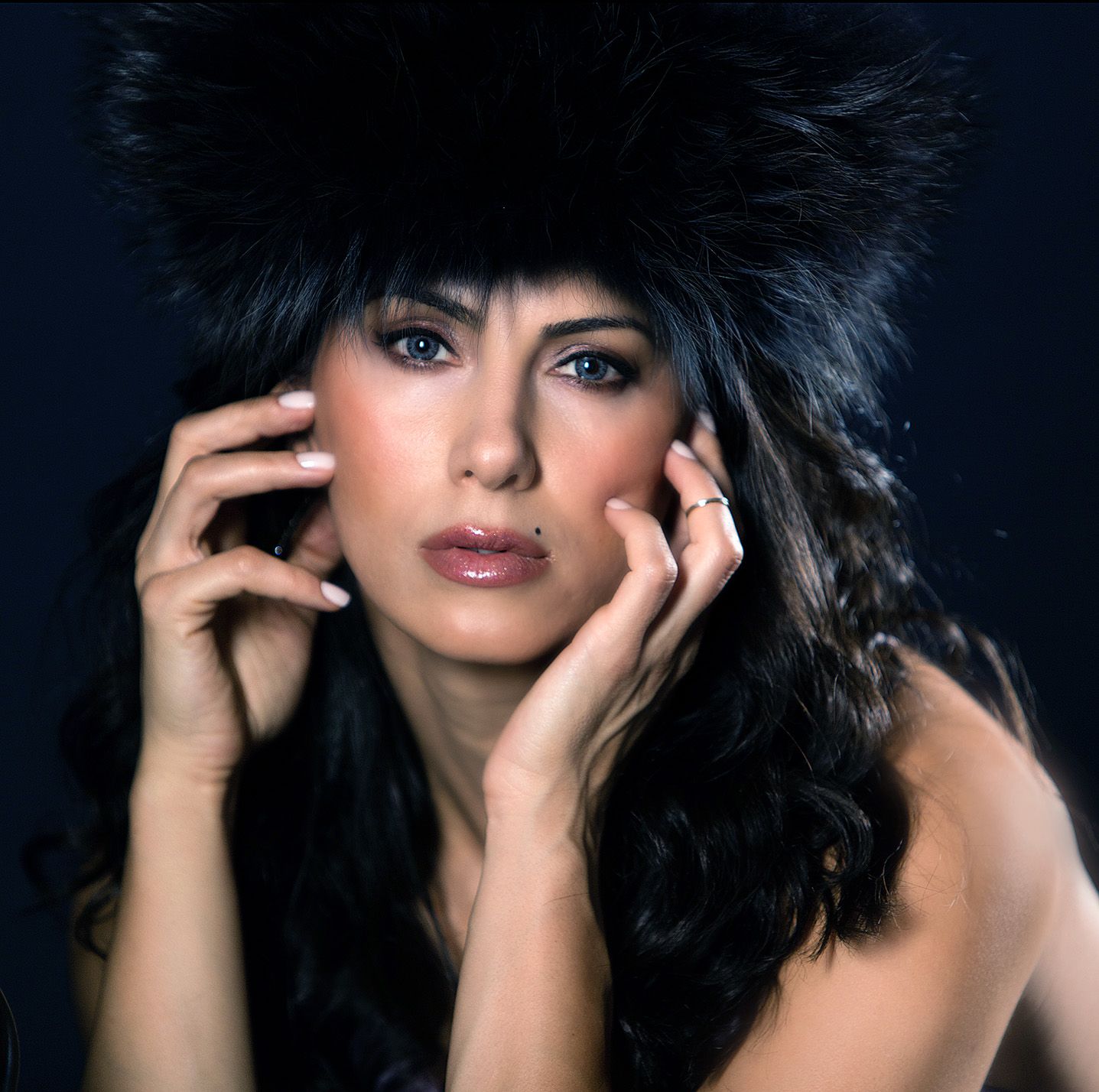A glamorous woman wearing a black fur hat poses elegantly with hands framing her face against a dark background.