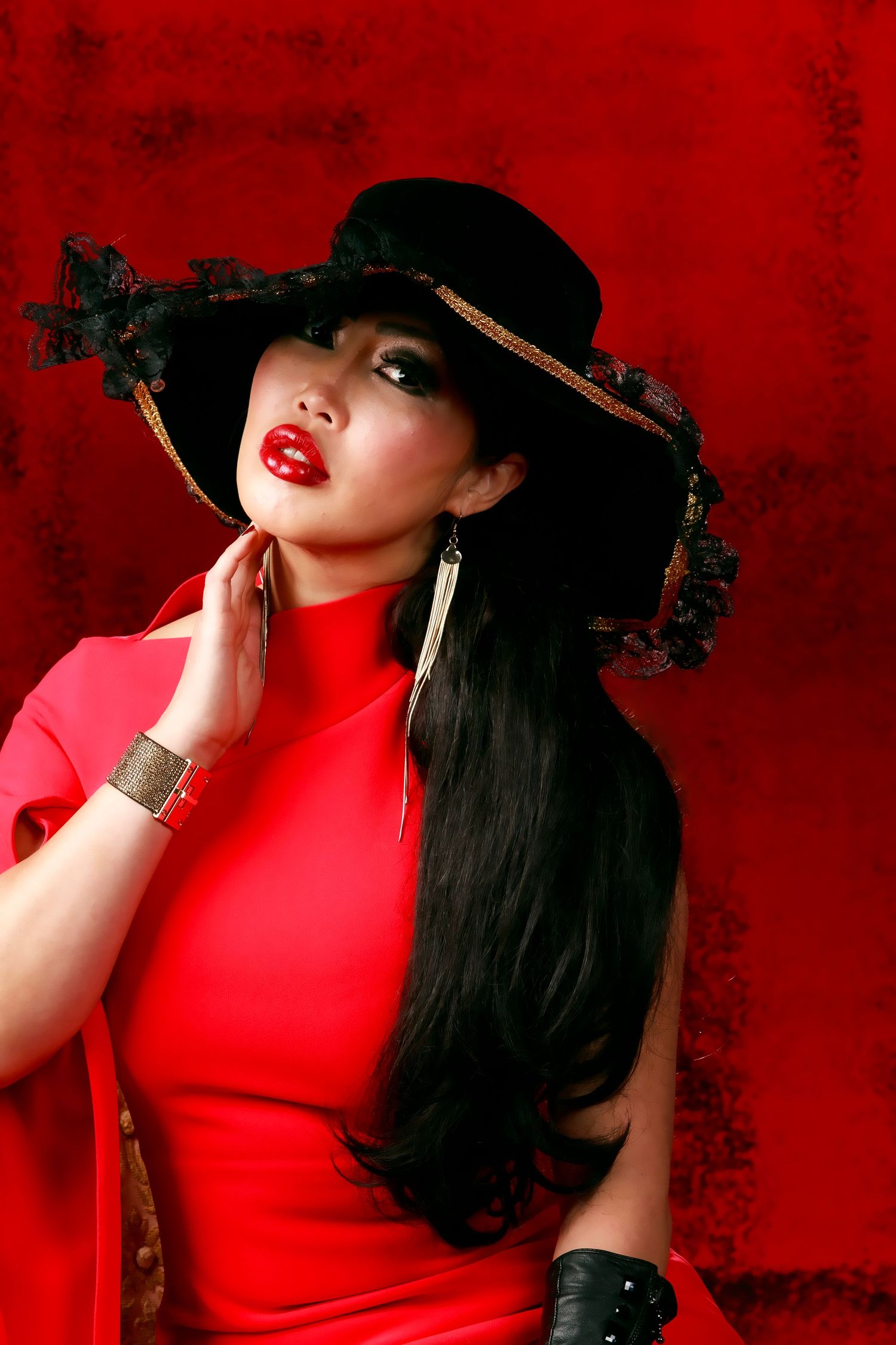 A glamorous woman in a red dress and black wide-brim hat with lace, wearing bold red lipstick against a red background.