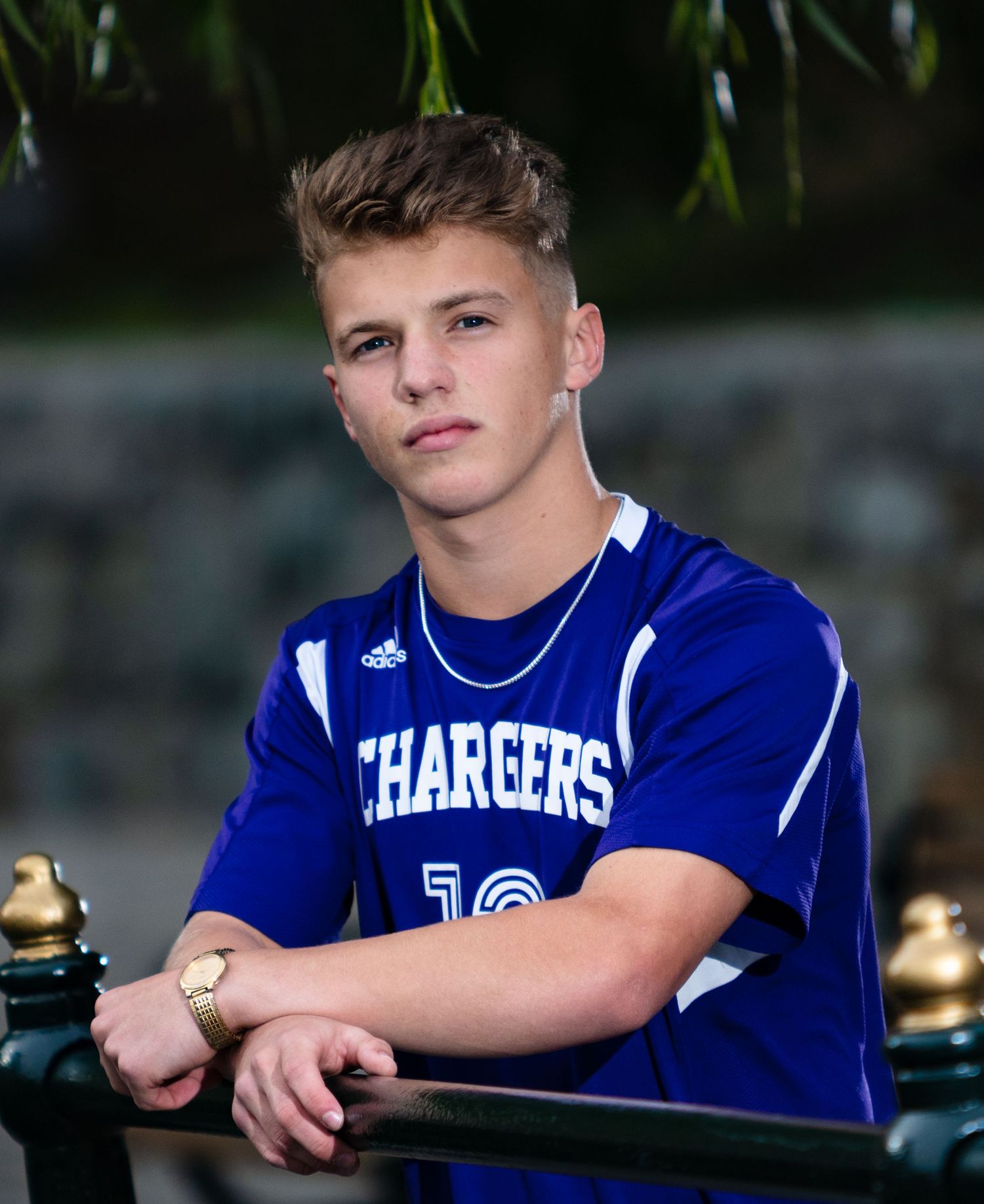 Teen boy in blue Chargers soccer jersey leaning on railing in outdoor senior portrait photo.