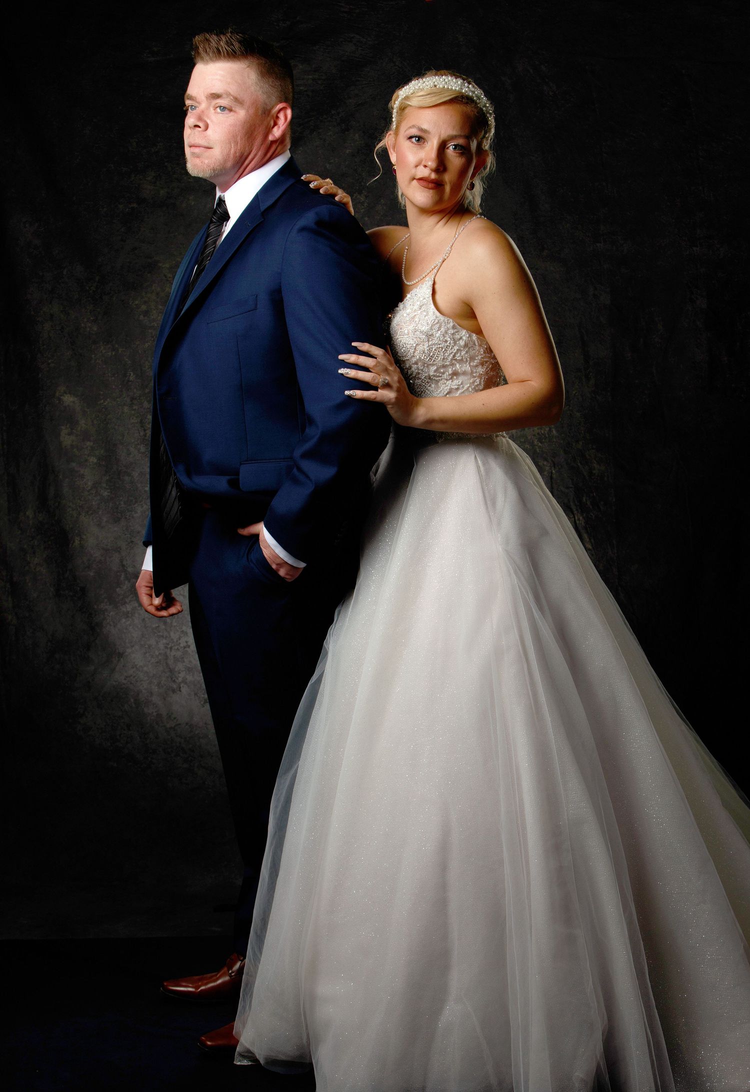 Bride in white ball gown and groom in navy suit pose back-to-back in a dramatic studio wedding portrait.