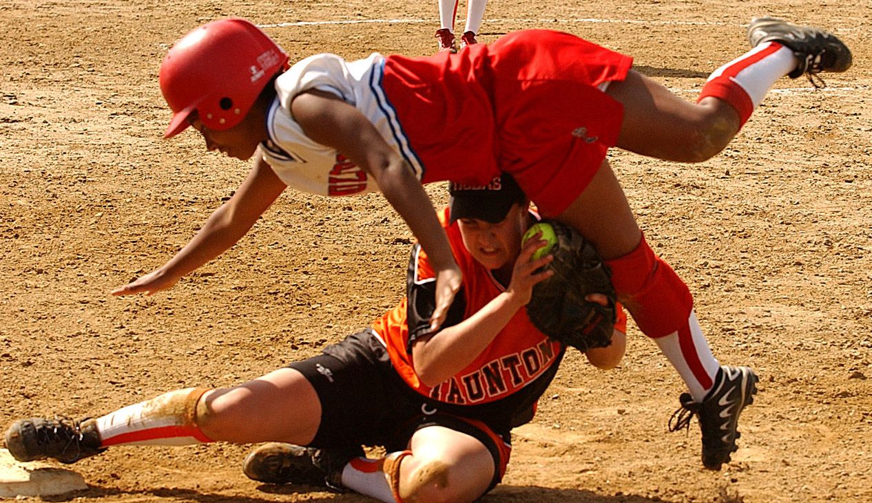 Two softball players collide at base during intense game, with runner sliding and fielder attempting tag on dirt field.