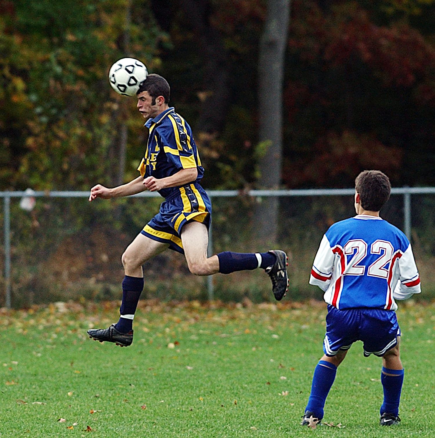 Soccer player in blue and yellow jersey heading the ball mid-air during a match, with opponent wearing number 22 nearby.