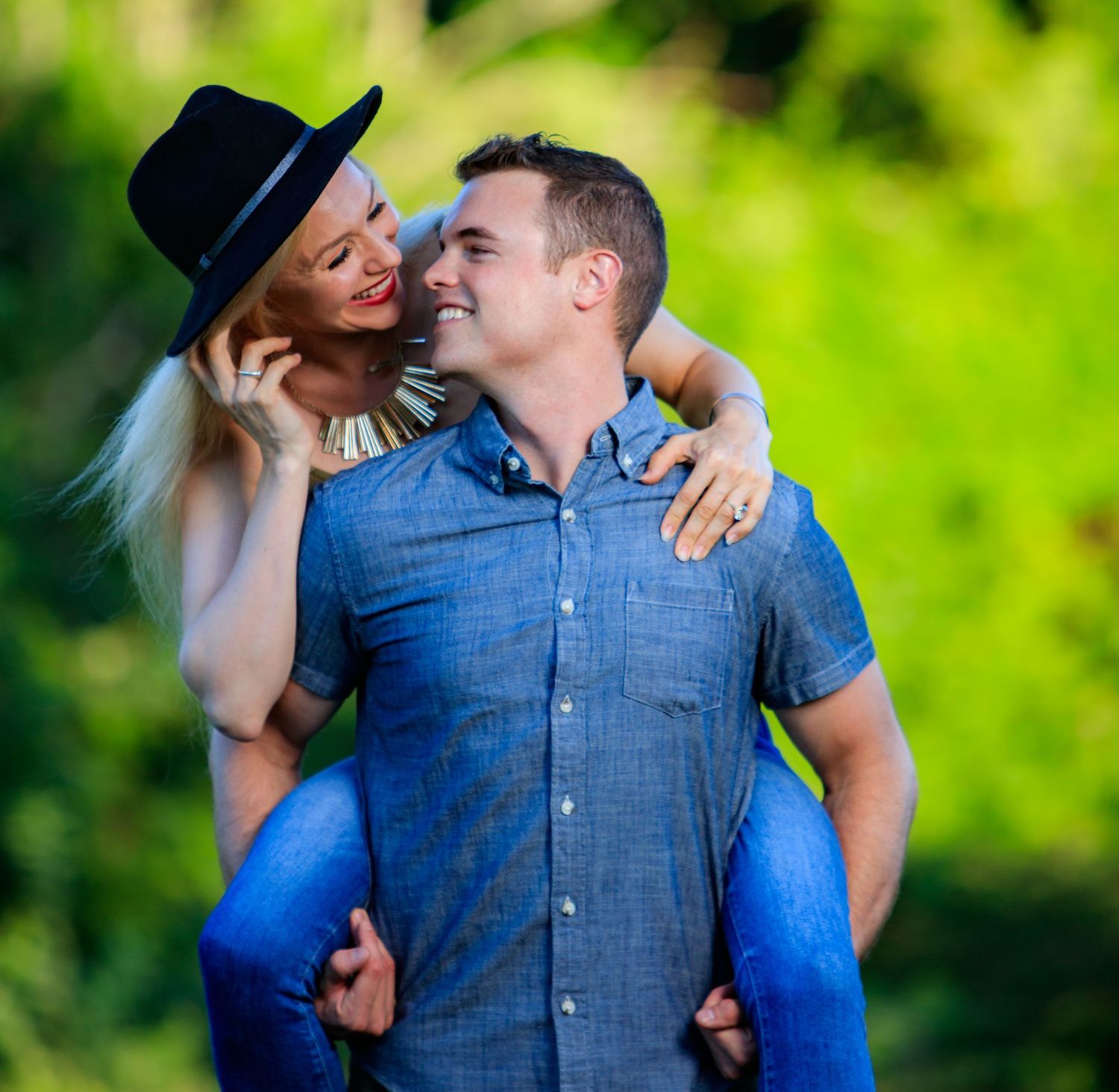 A smiling blonde woman in a black hat rides piggyback on a happy man in a blue shirt outdoors.