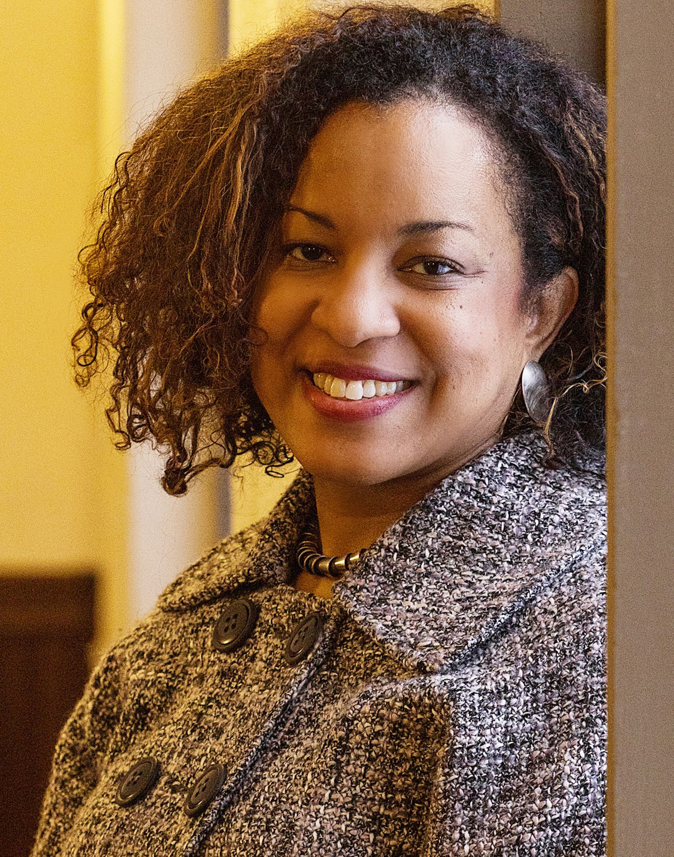 Smiling woman with curly hair wearing a gray tweed jacket and earrings, leaning against a wall indoors.