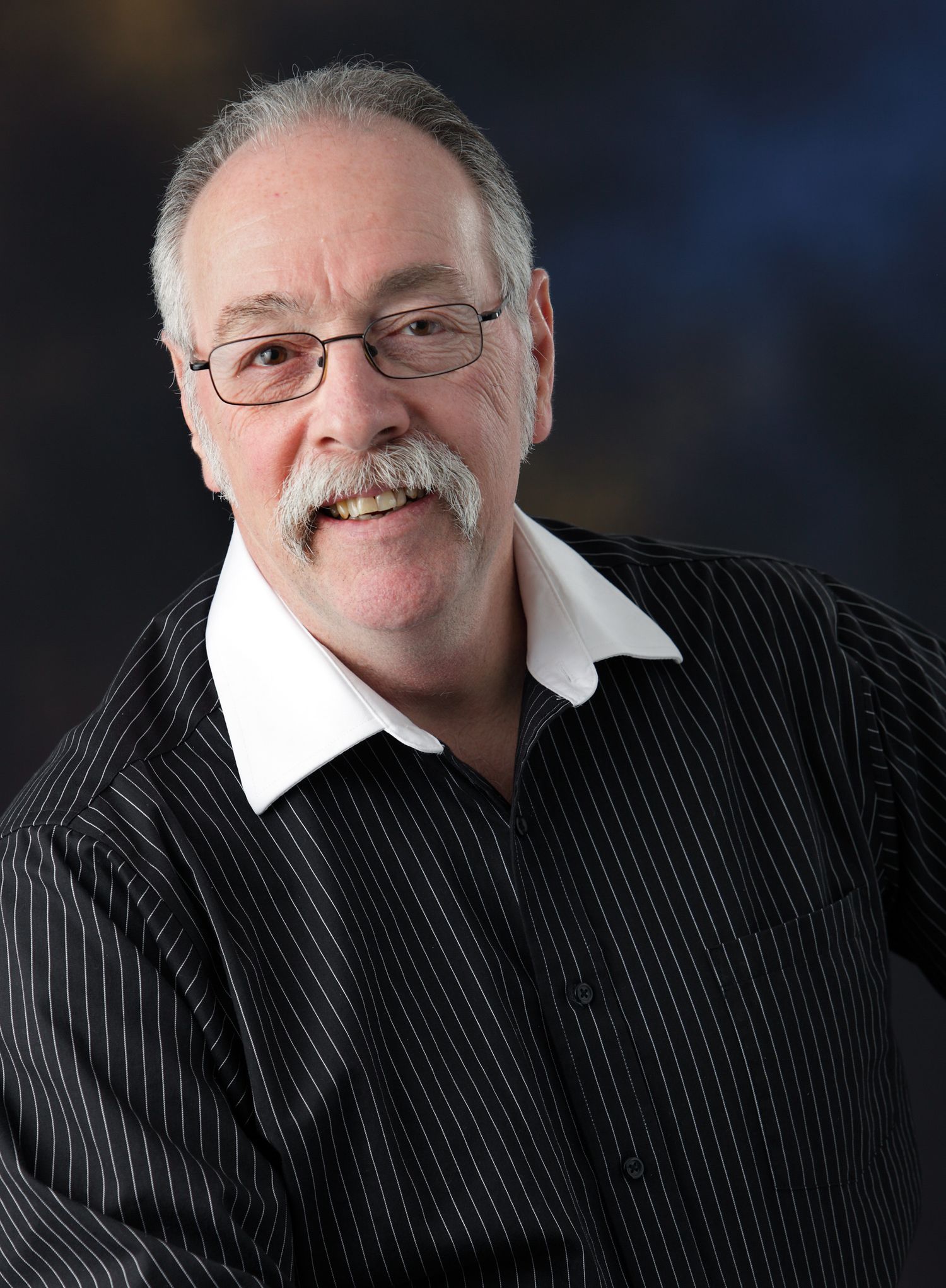 Senior man with gray mustache and glasses in a black pinstripe shirt posing for a professional headshot photo.
