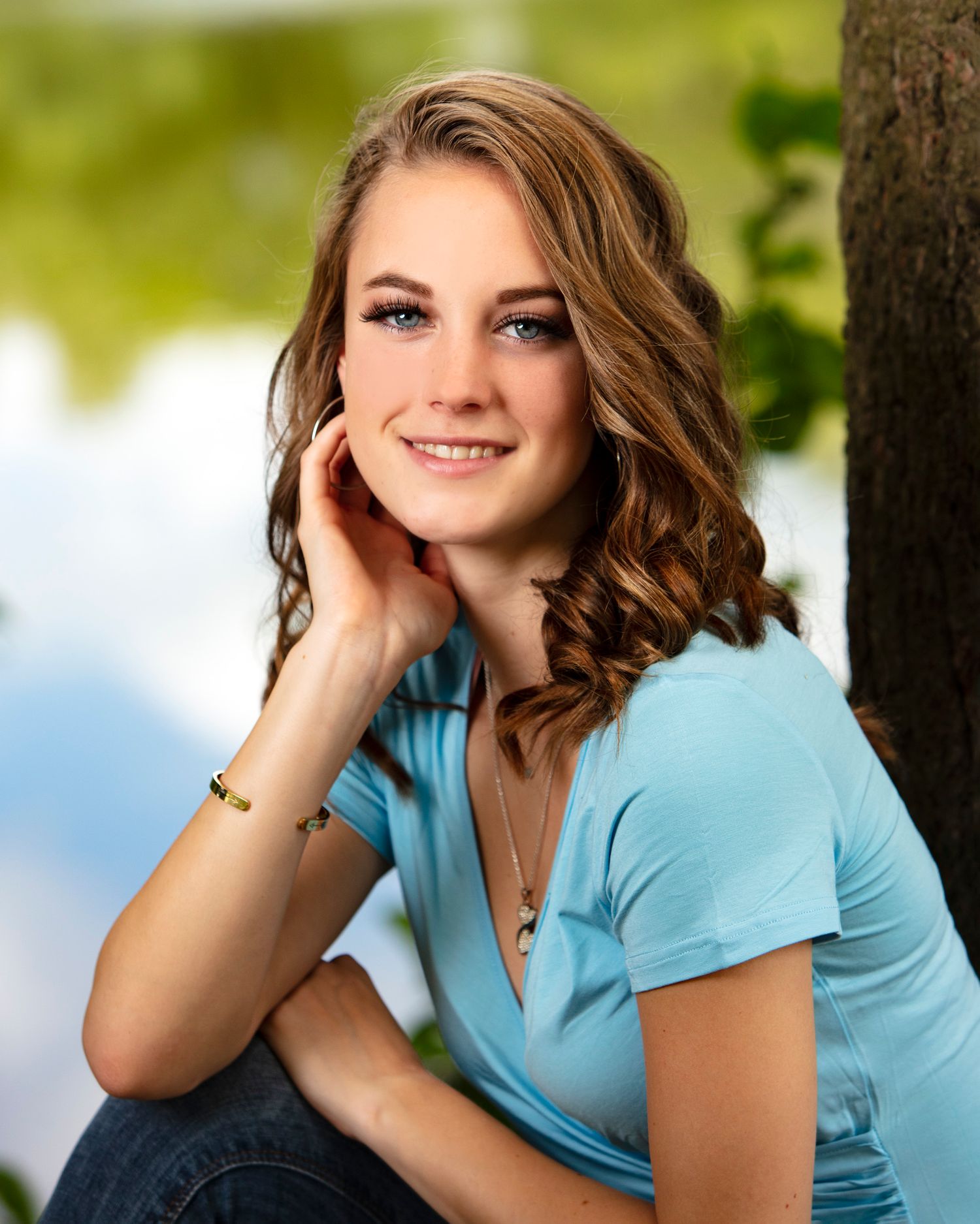 Smiling young woman with curly auburn hair in blue top sitting by tree outdoors.