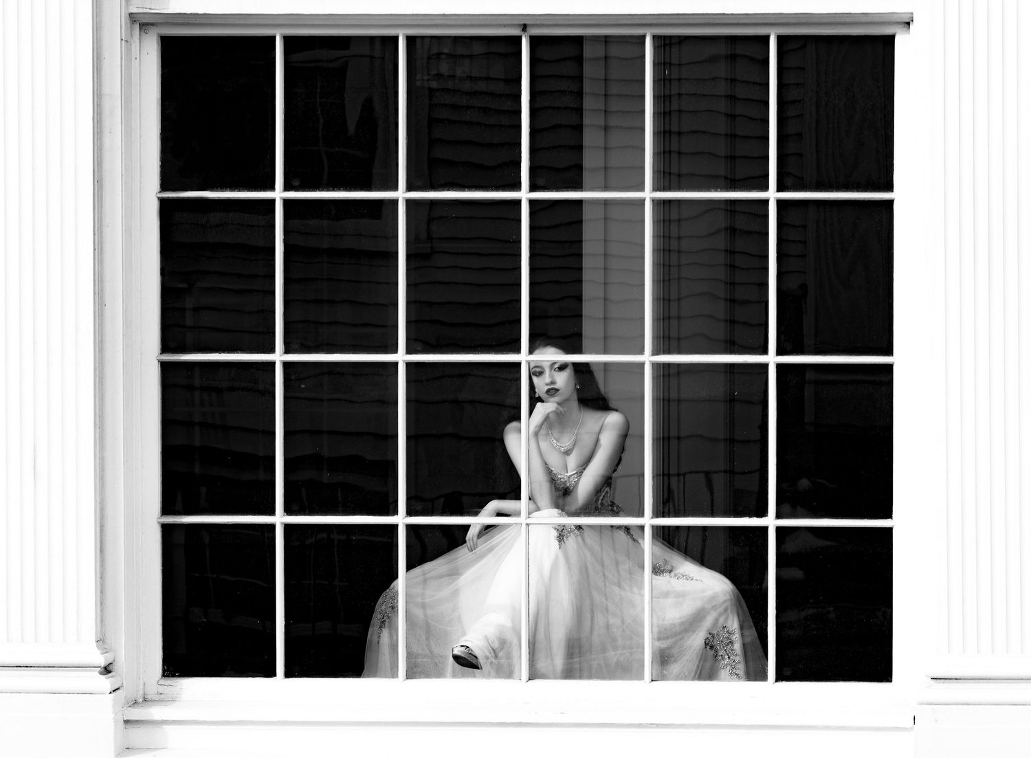 Black and white photo of a bride in a gown posing behind a large window with grid panes.