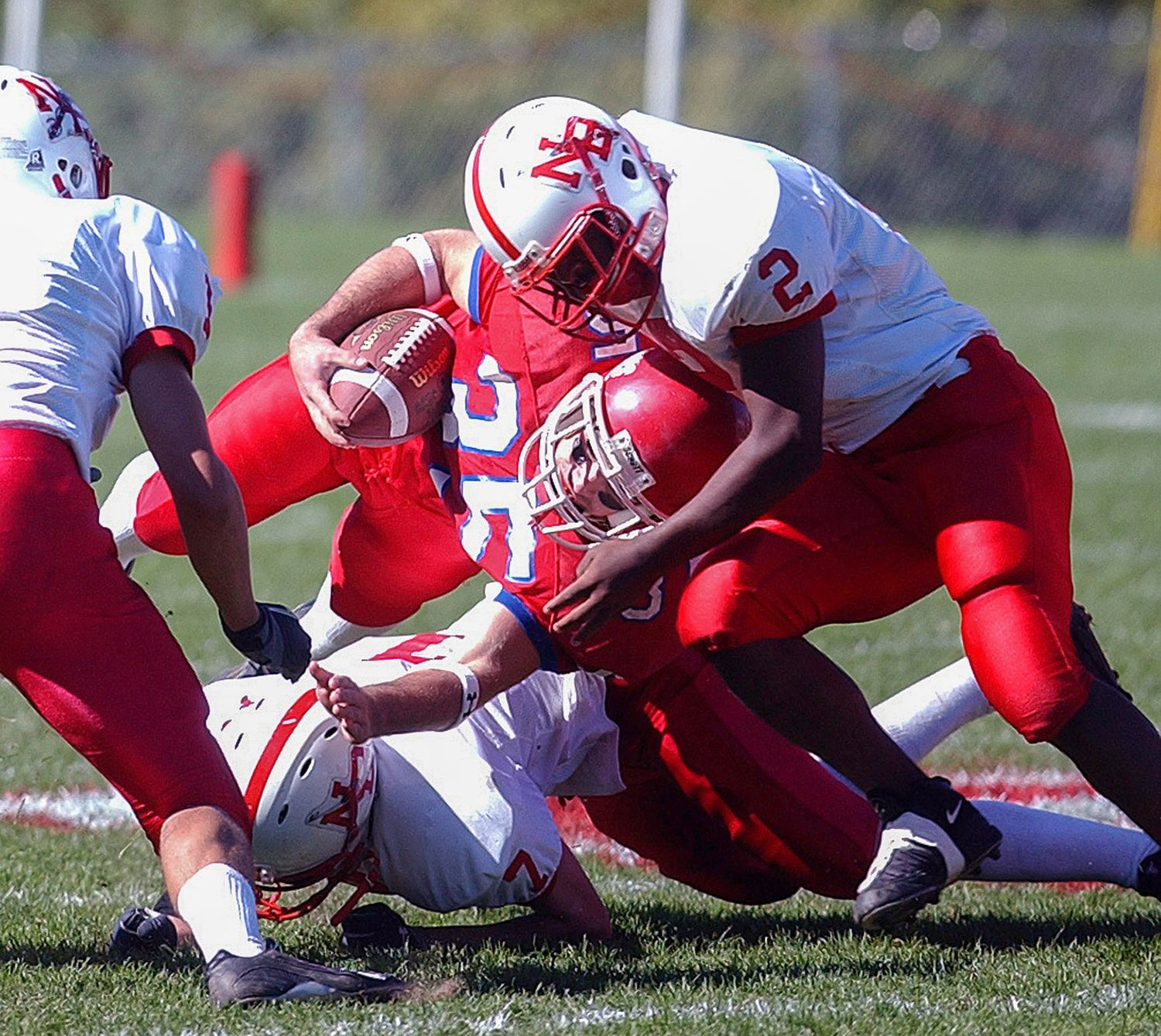 Red team football players tackle ball carrier during intense high school football game on sunny day.