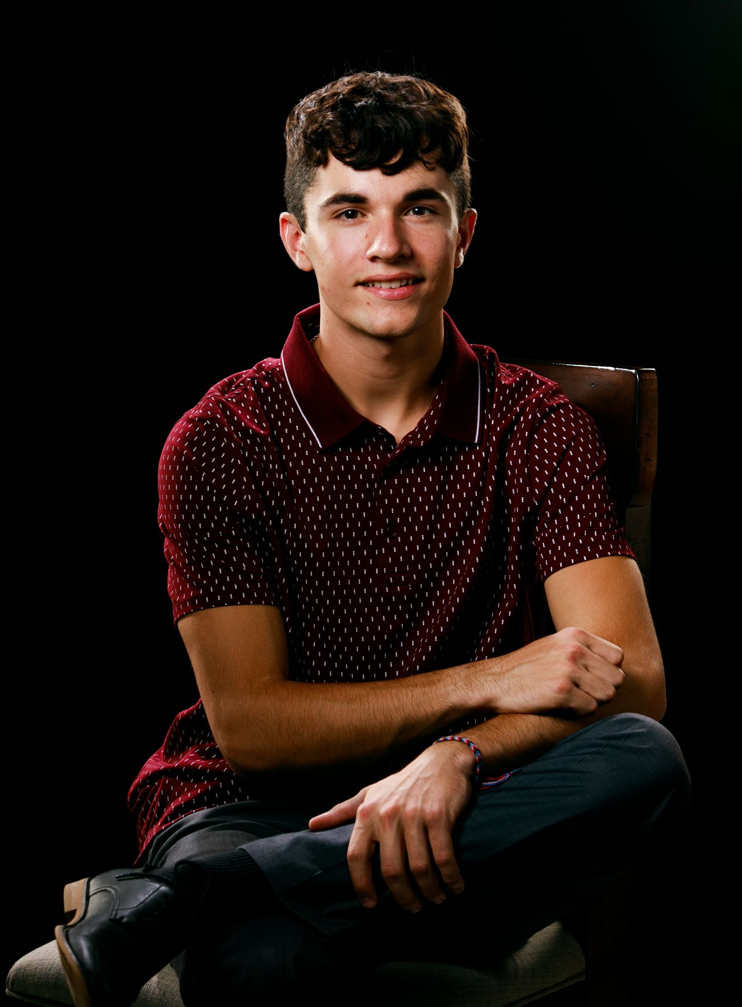 Smiling teenage boy in red polo shirt sits cross-legged in chair for senior portrait.