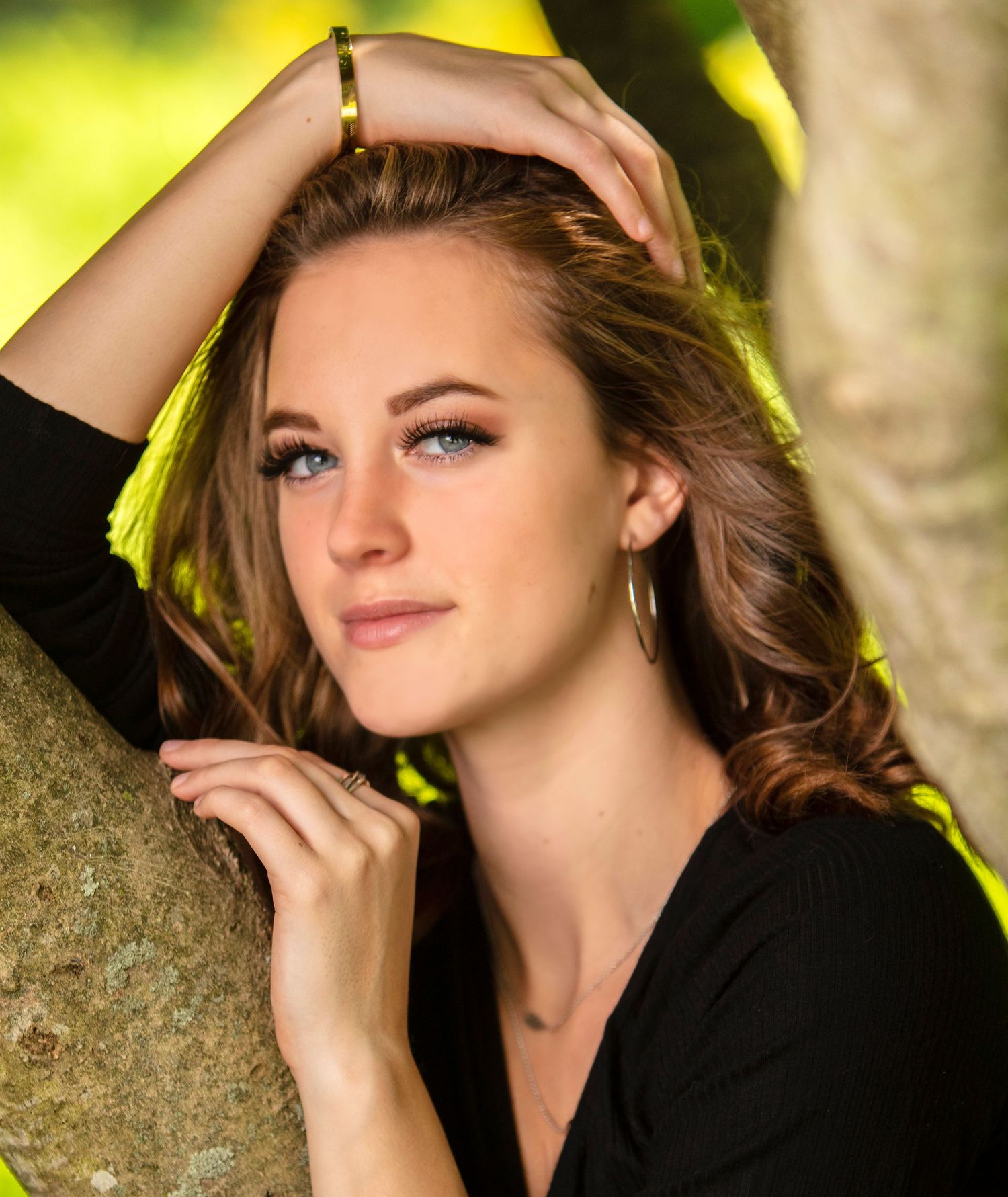 Young woman with blue eyes and brown hair posing against tree in black top.