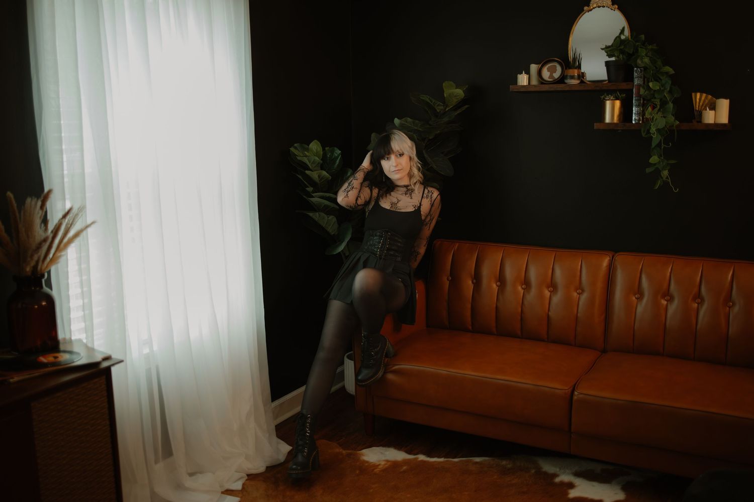 Woman in black outfit posing by leather sofa near window in dark moody interior.