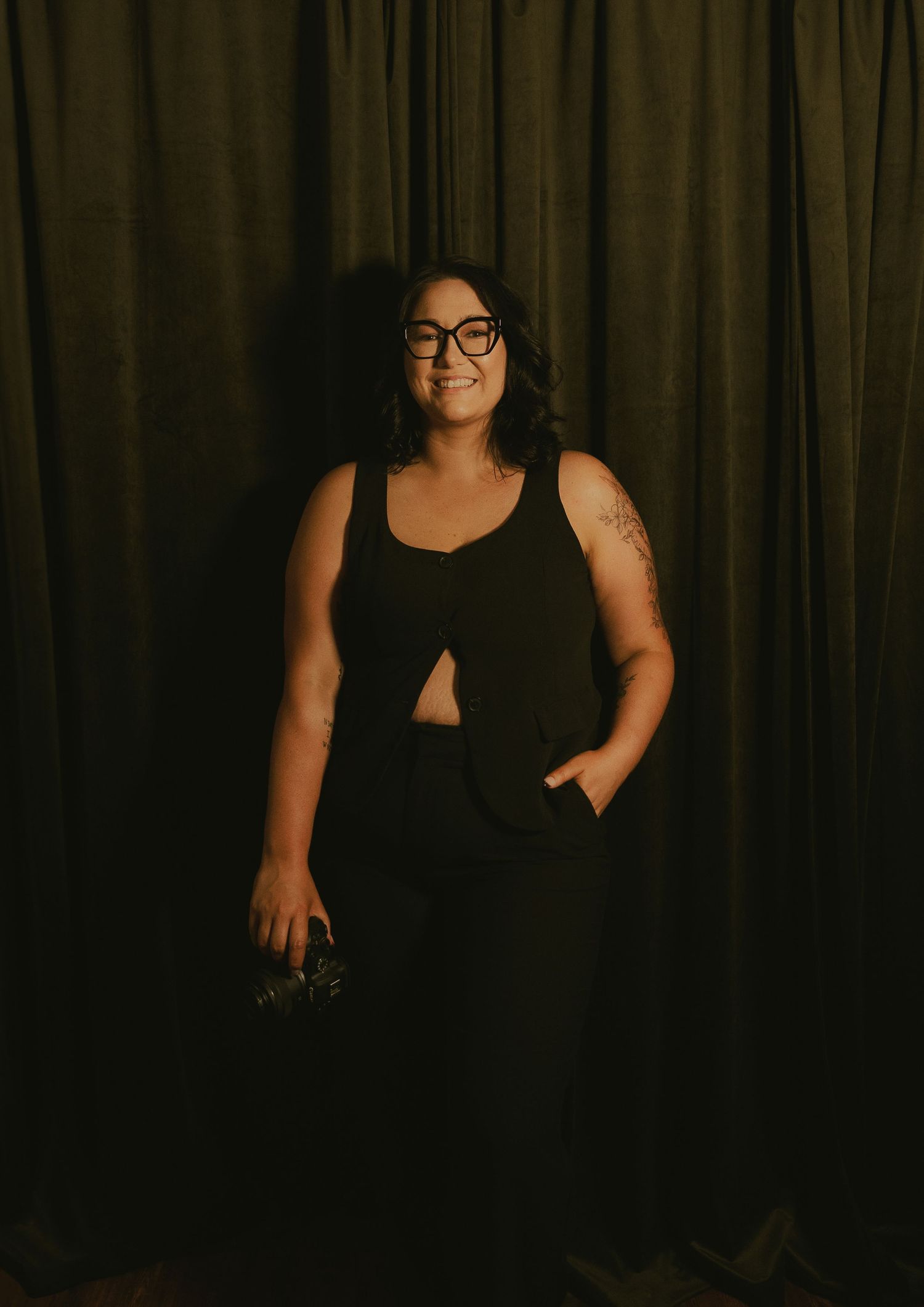 Woman in black outfit with glasses smiling against dark curtain backdrop.