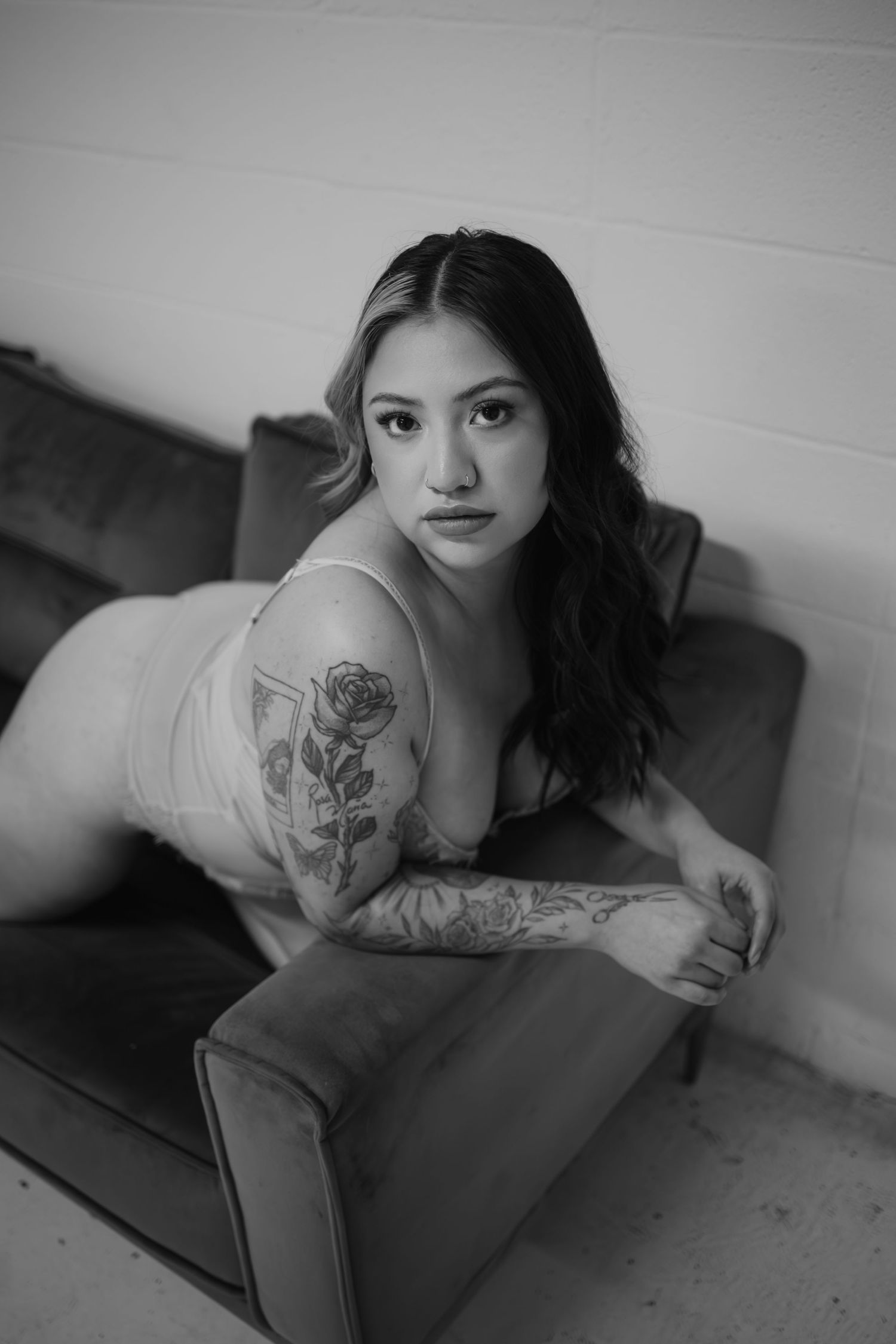 Black and white boudoir portrait of tattooed woman in sheer lingerie on velvet chair.