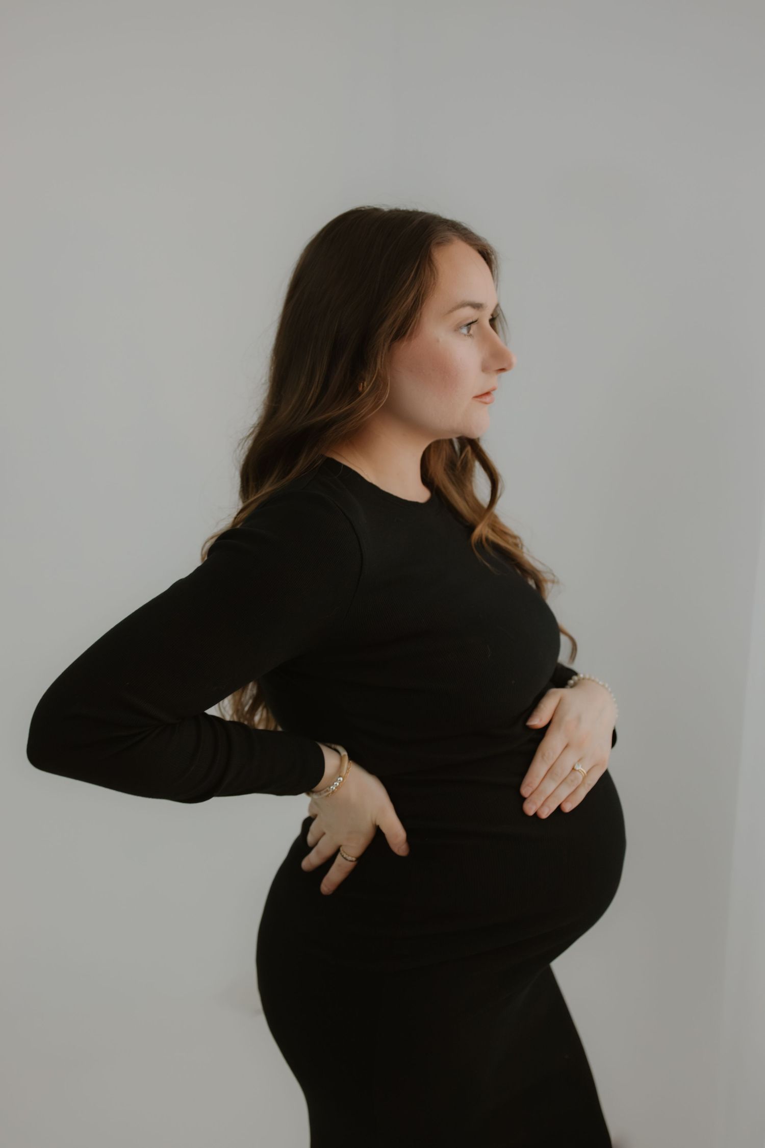 Pregnant woman in black dress cradling baby bump in profile pose against white background.