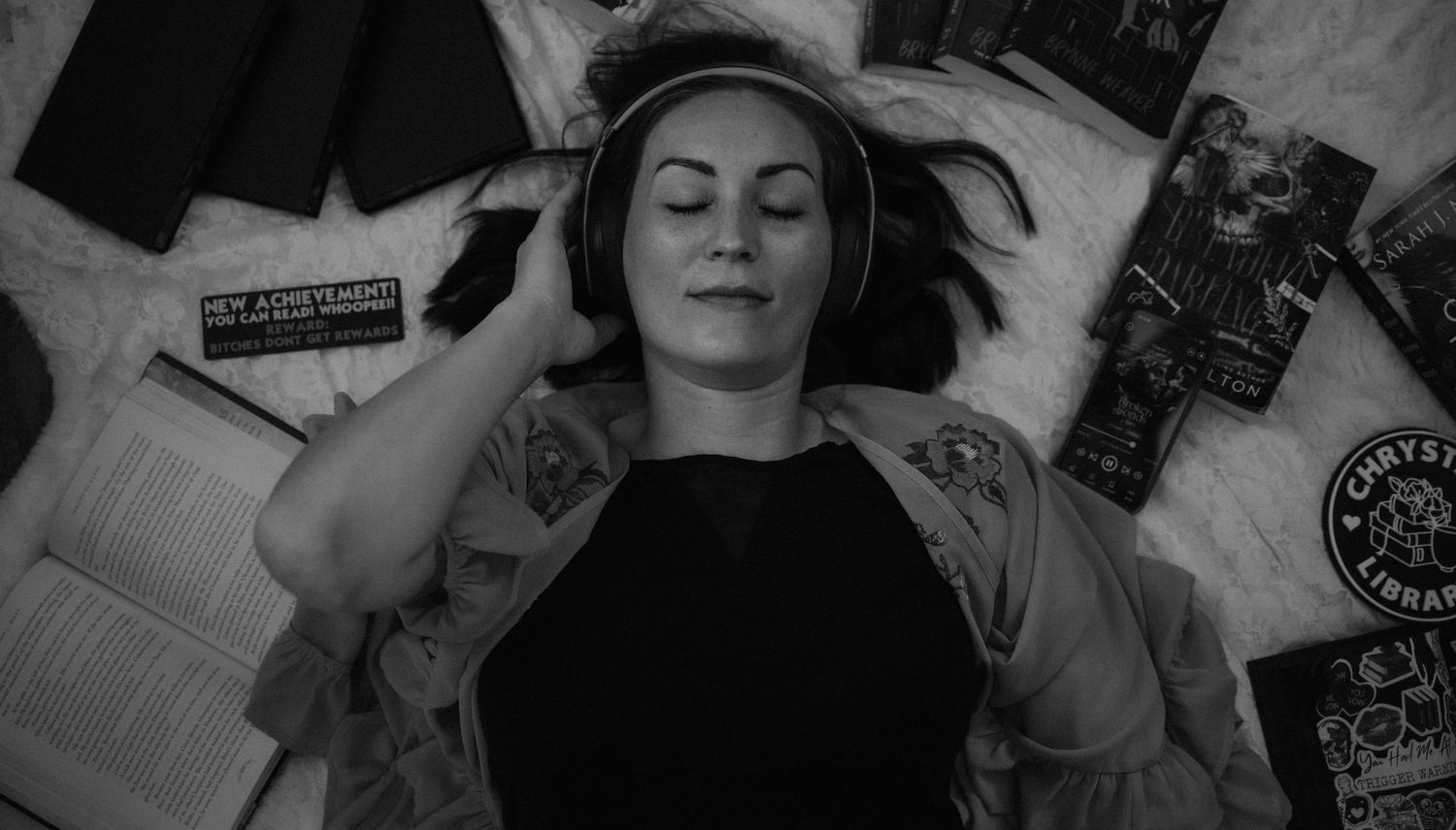Black and white photo of tattooed woman lying among books listening to headphones peacefully.
