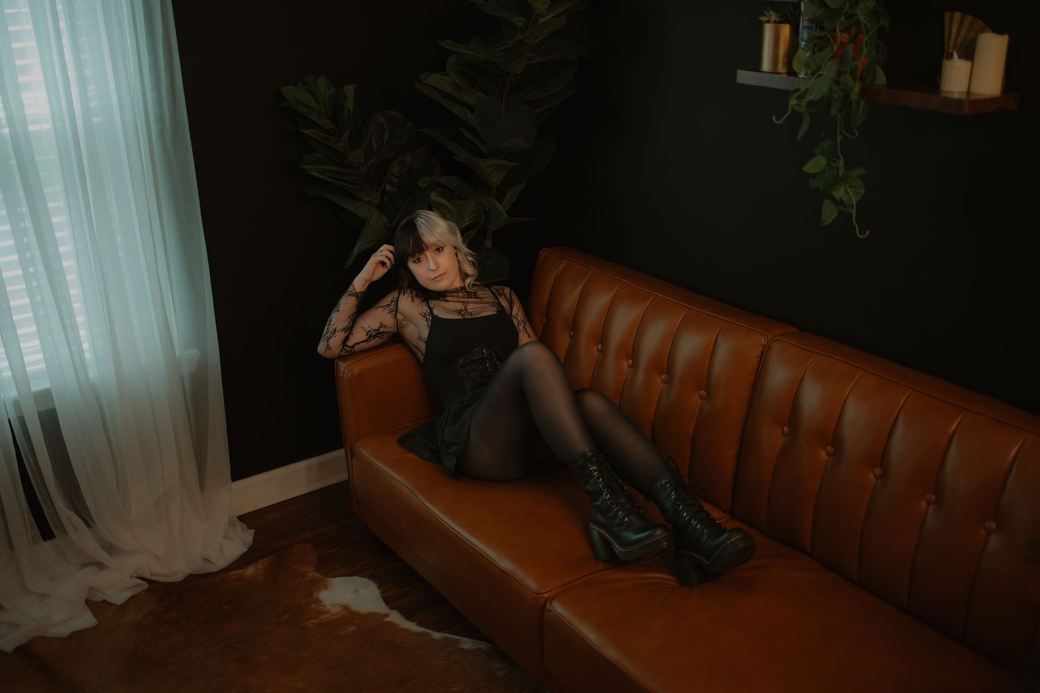 Tattooed woman in black slip dress and platform boots posing on a brown leather sofa.