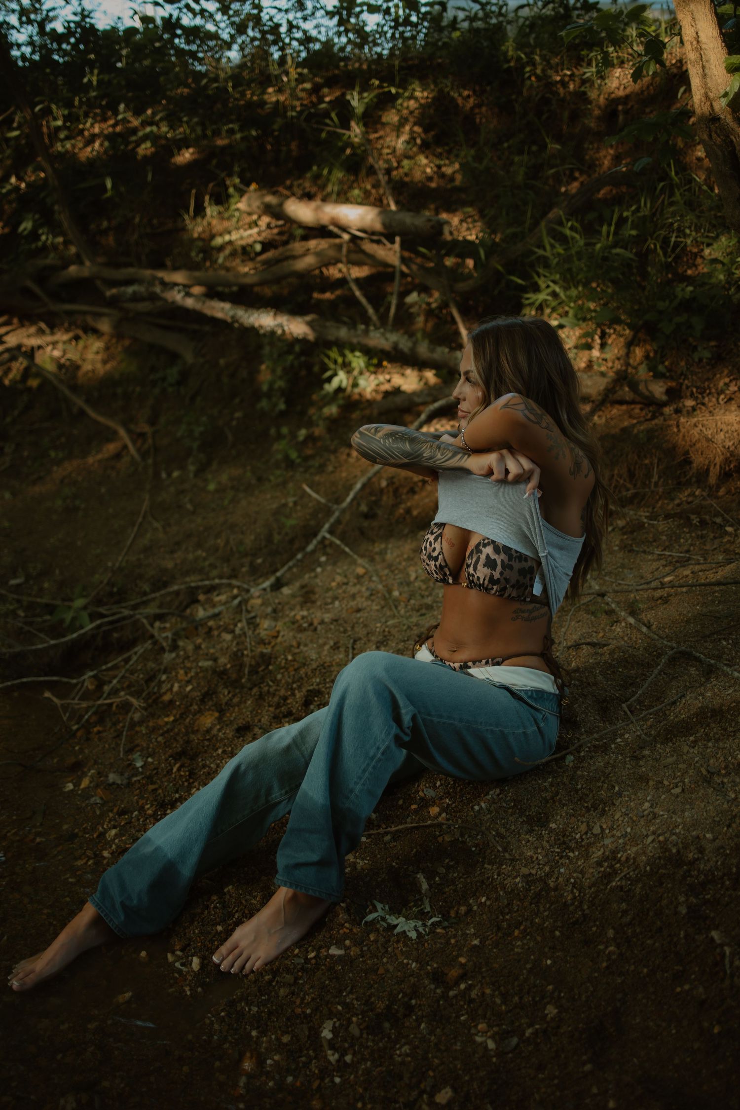 Tattooed woman in crop top and jeans sitting barefoot on a hillside in natural surroundings.