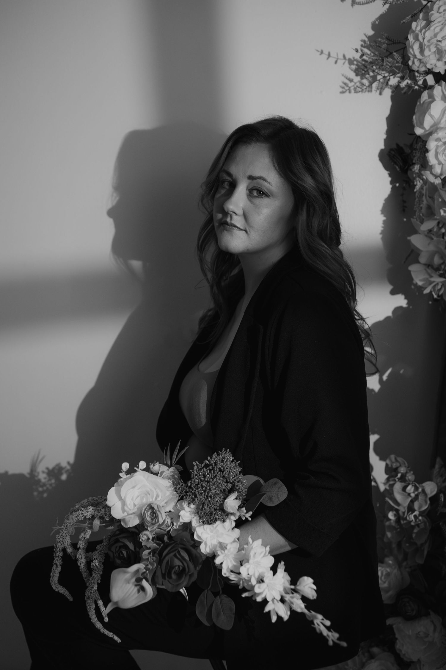 Black and white portrait of woman holding floral bouquet leaning against wall with dramatic shadow.