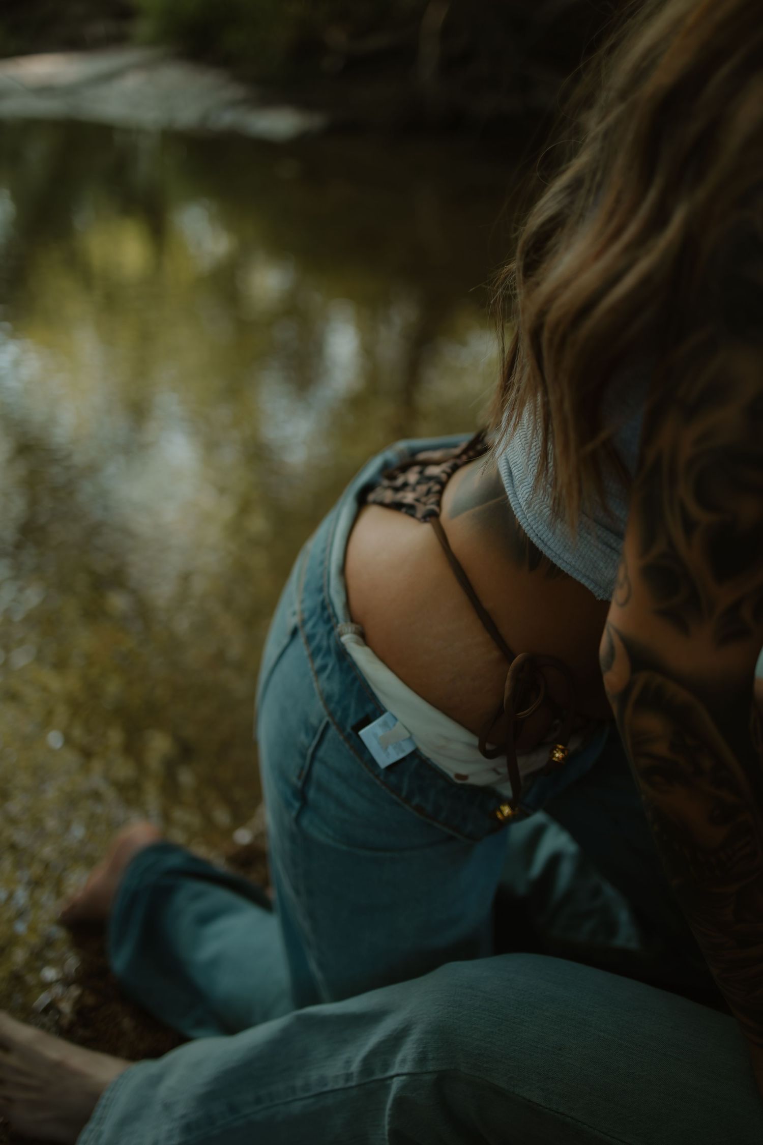 Close-up of tattooed woman in ripped jeans sitting by a reflective creek in nature.