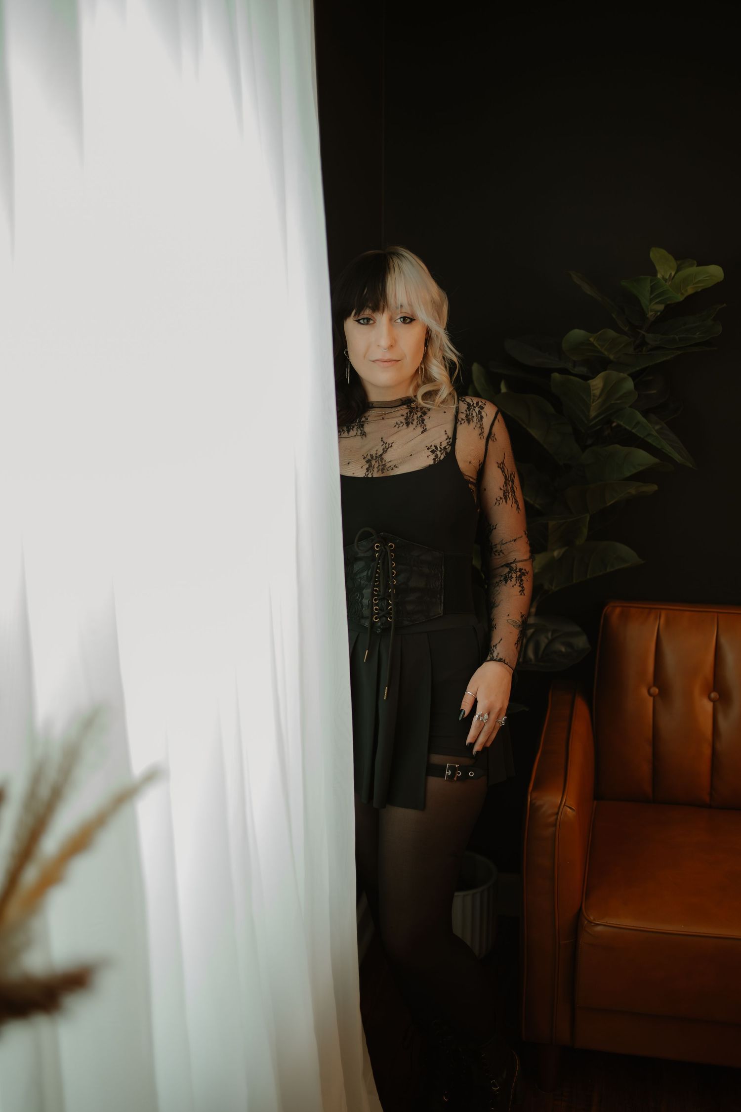 Woman in black lace bodysuit and corset peers around white curtain in moody dark studio.