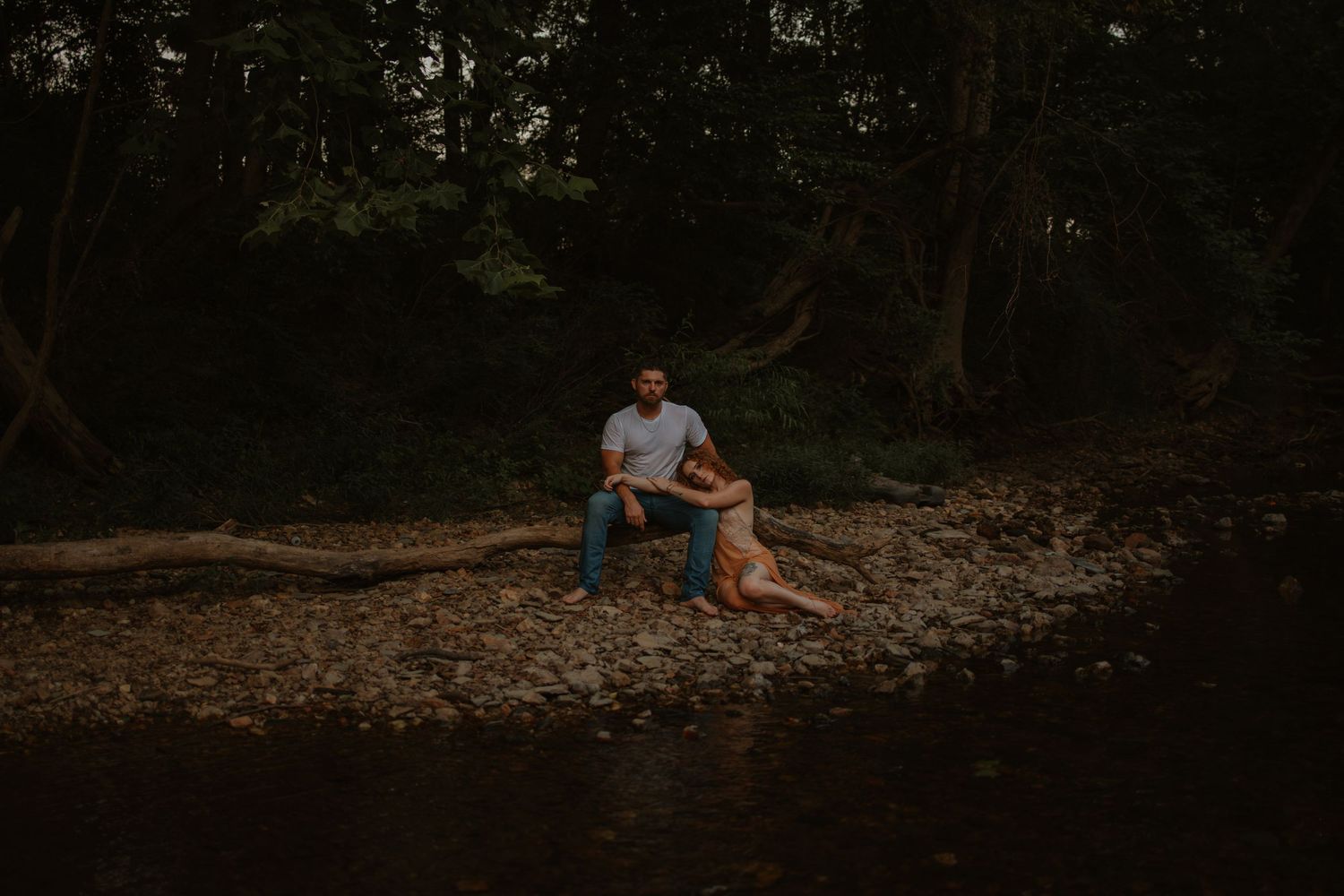 Couple sits together on rocky riverbank surrounded by dark lush forest at dusk.