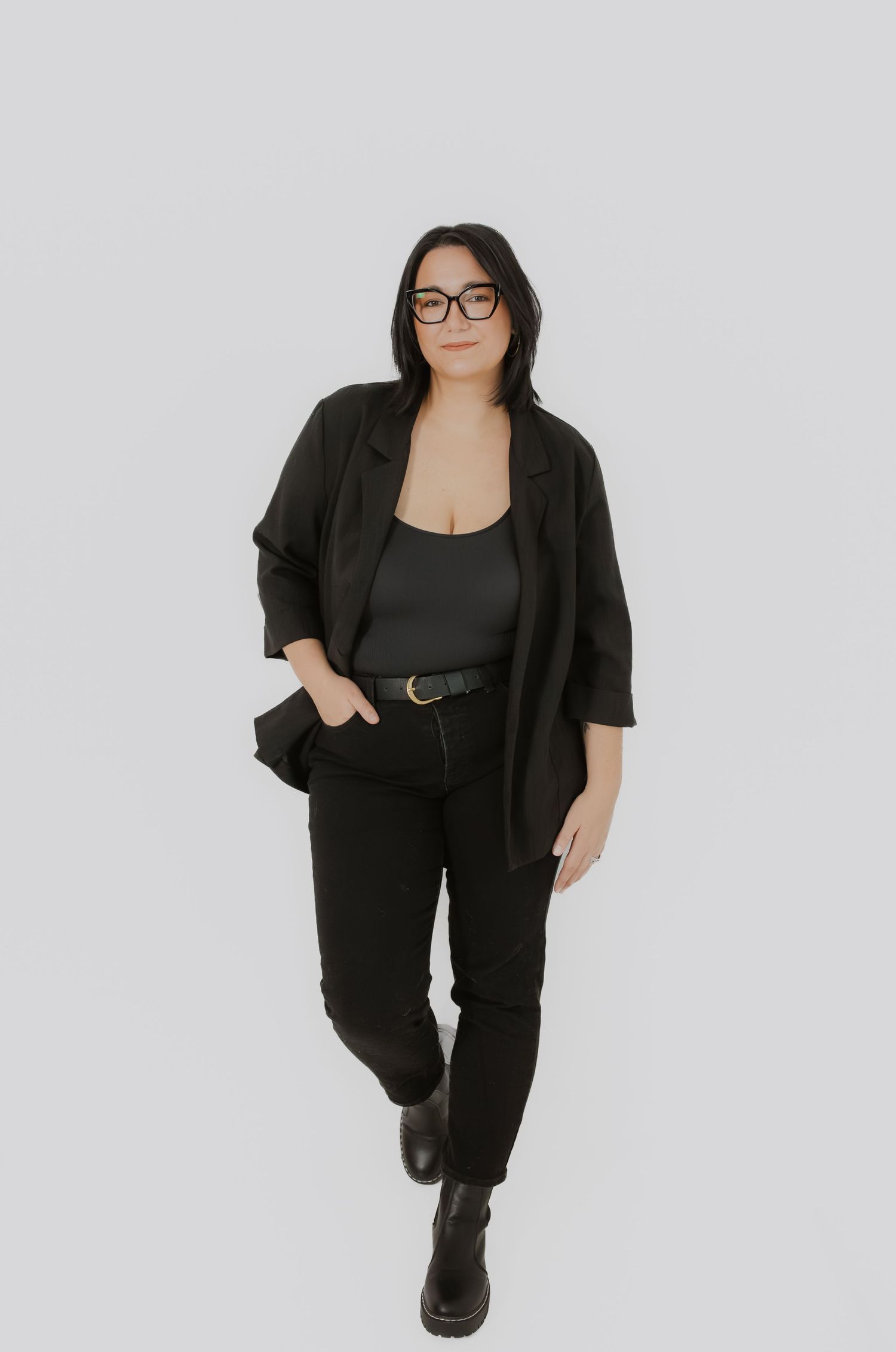 Plus-size woman in black blazer, jeans, and boots posing confidently on a white background.