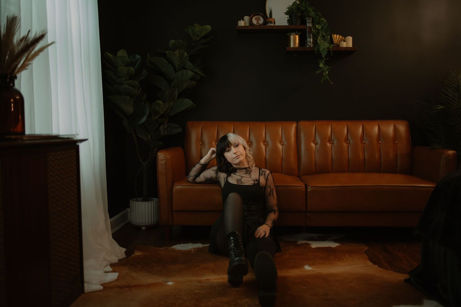 Young woman with tattoos sitting on floor in dark moody living room with leather couch and plants.