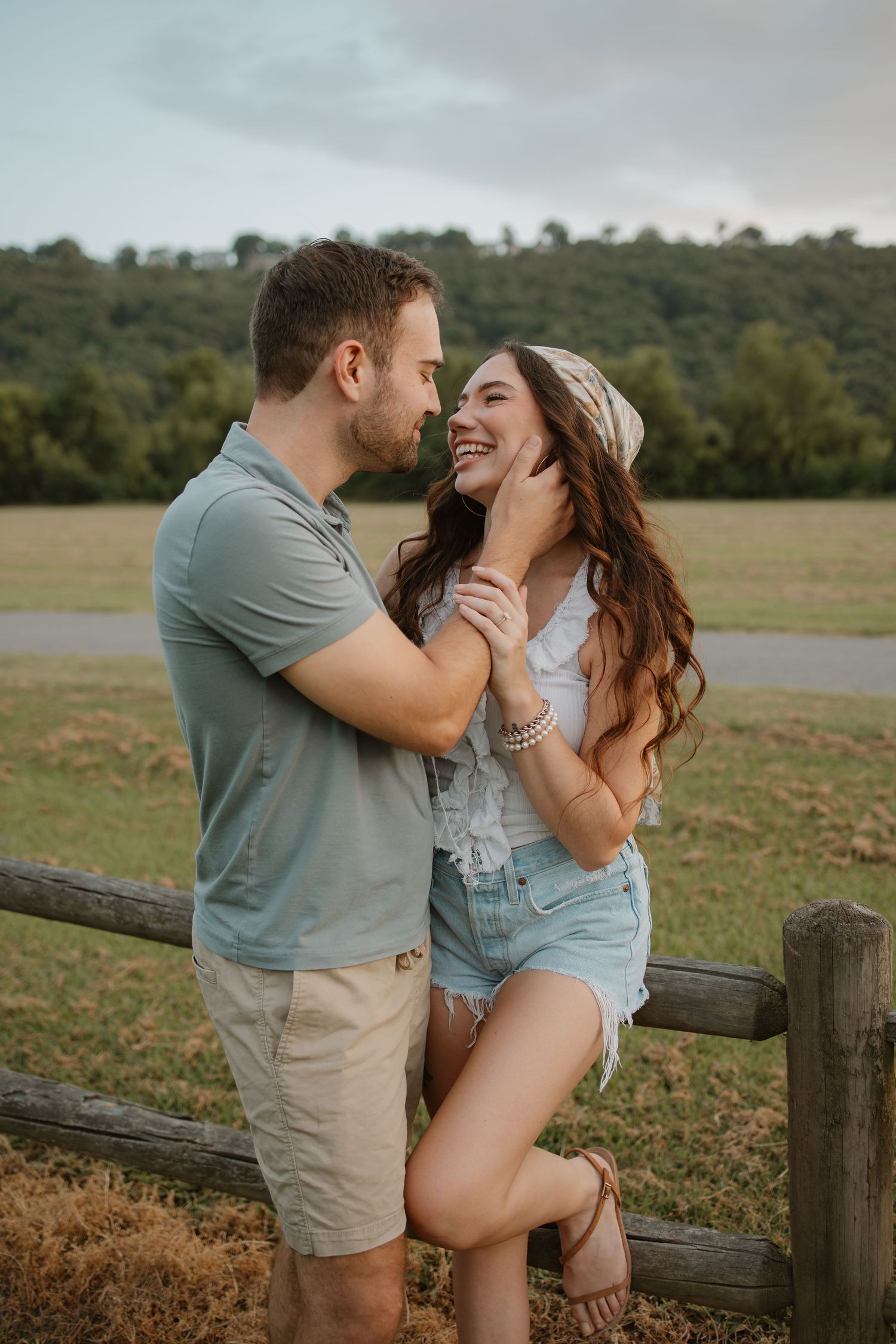 Couple sharing a romantic moment by a rustic fence in an open field, woman laughing as man gently cups her face.