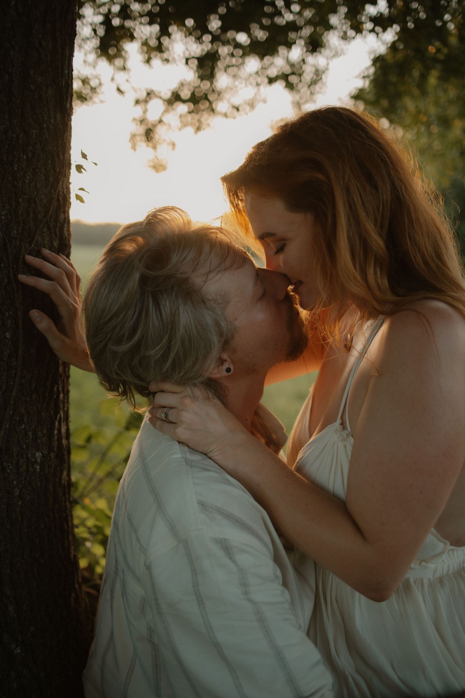Two women sharing a tender kiss near a tree in a sunlit outdoor setting at golden hour.