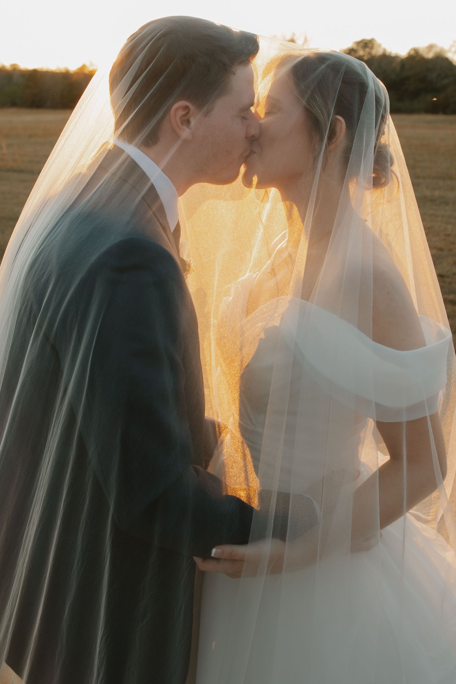 Bride and groom kissing under sheer veil at golden sunset in open countryside field.