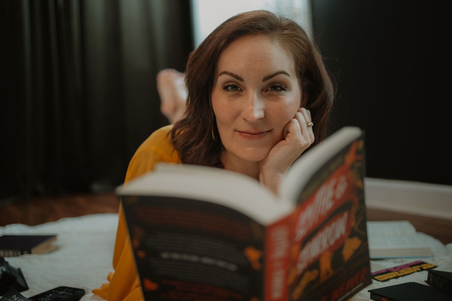 Woman lying down smiling while reading a book, captured in a warm candid lifestyle portrait.