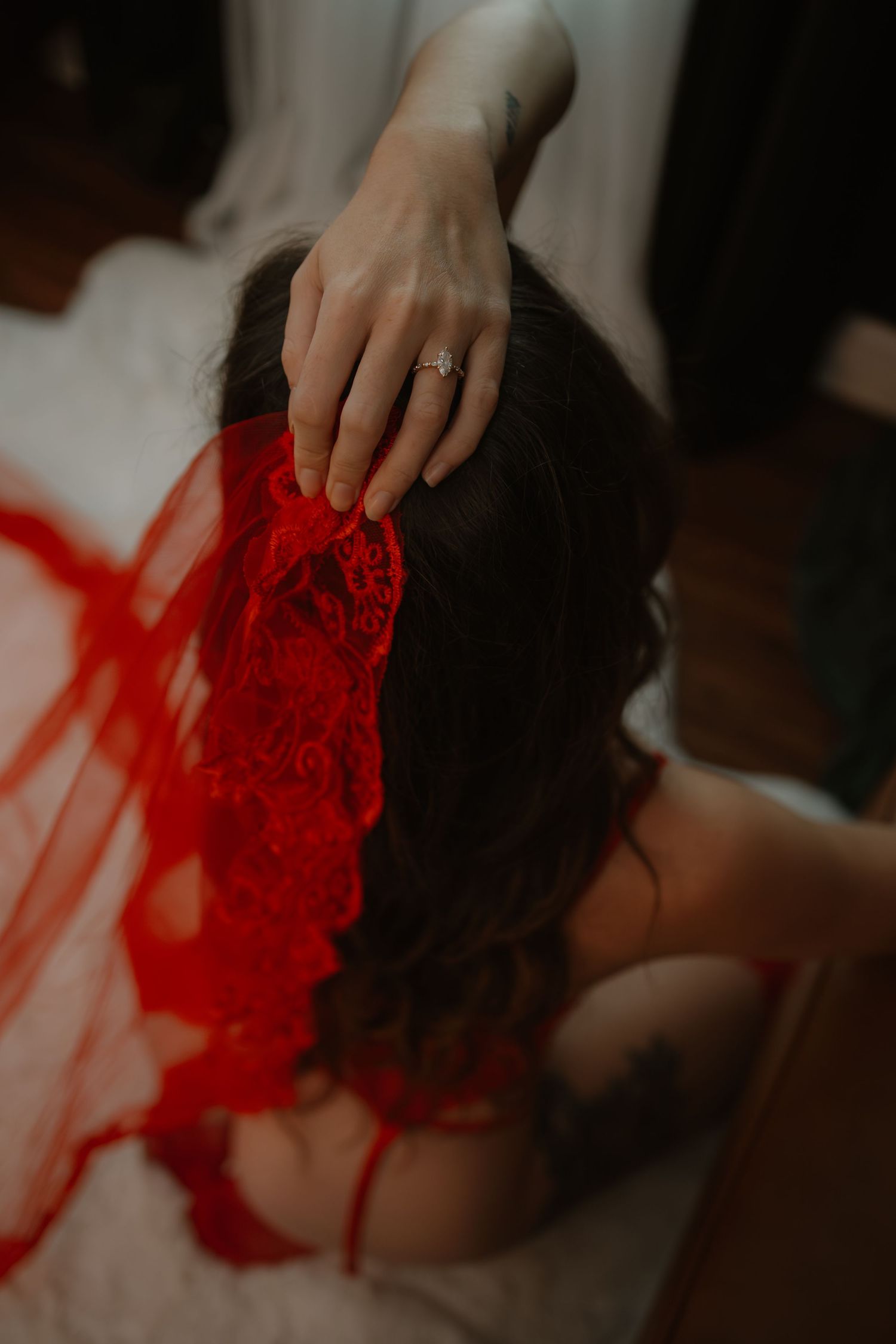 A woman with dark hair wearing red lace lingerie and a diamond ring rests her hand gently on her head.