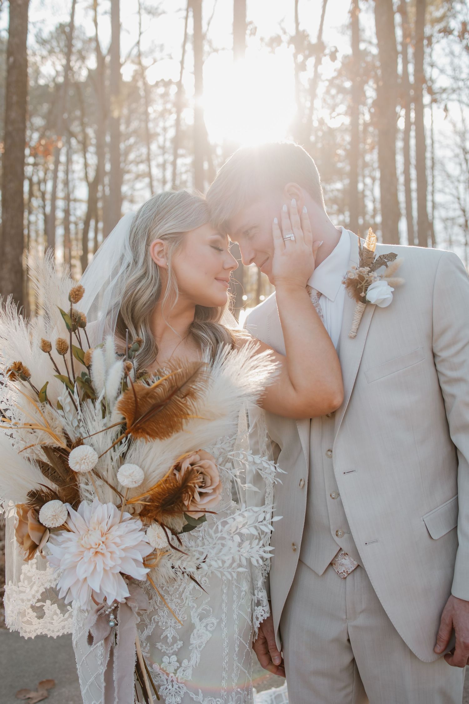 Bride and groom touching foreheads in sunlit forest with boho floral bouquet and veil.