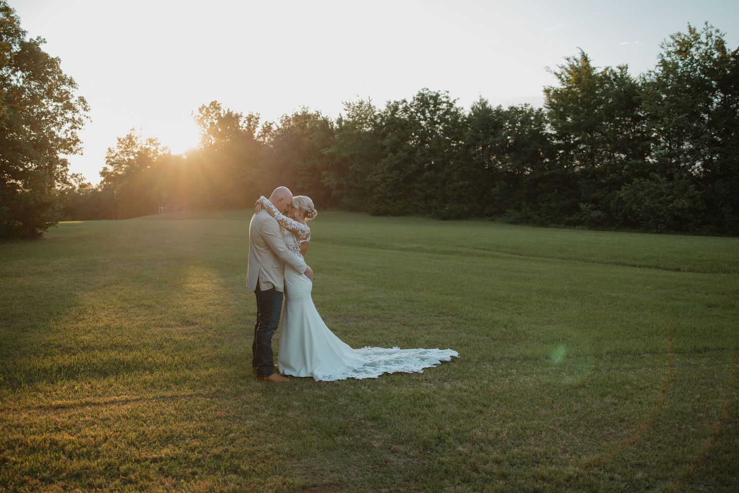 Bride and groom kissing at golden hour sunset in open green field with tree line backdrop.