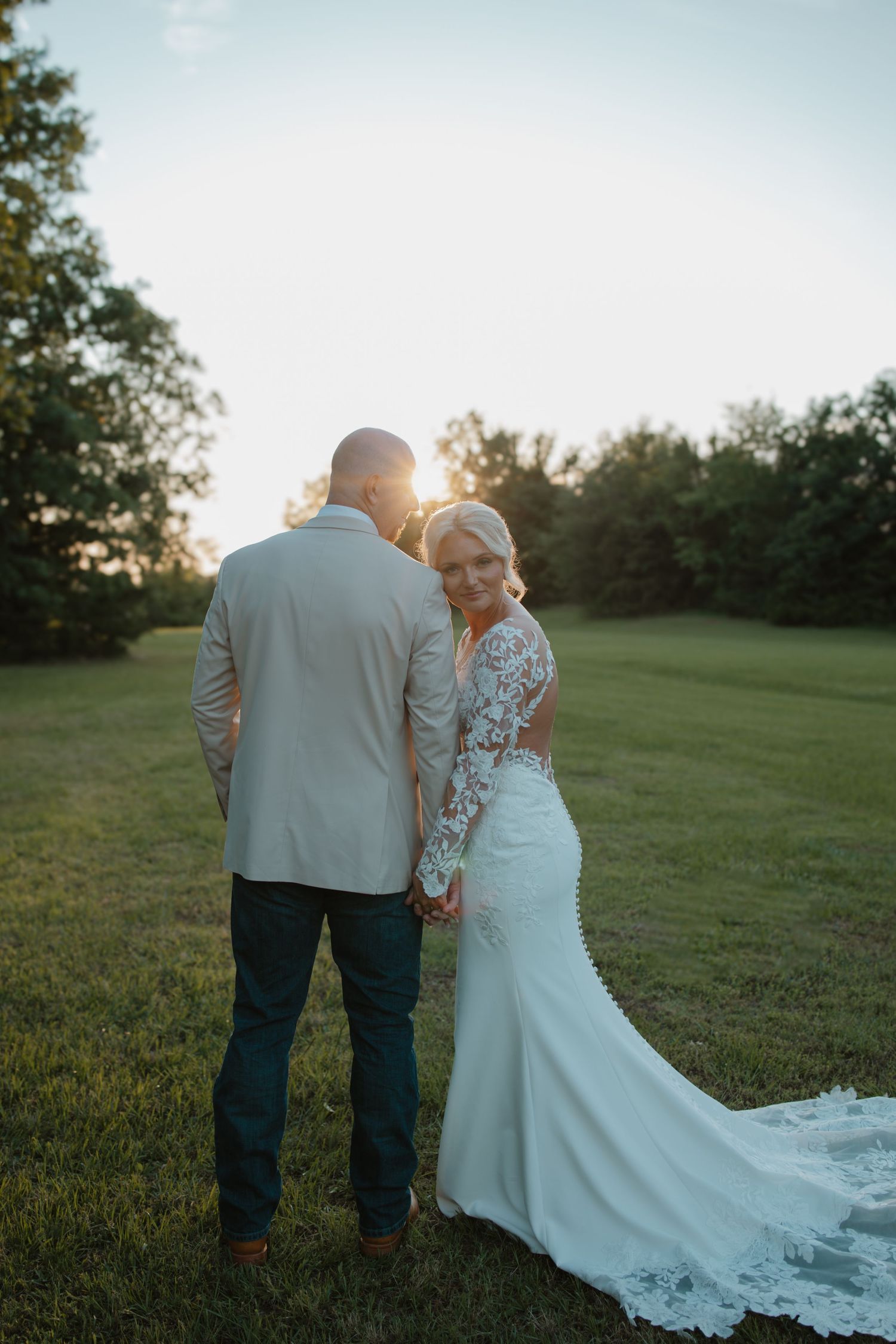 Bride in lace gown and groom in tan blazer pose at golden hour in a lush green field.