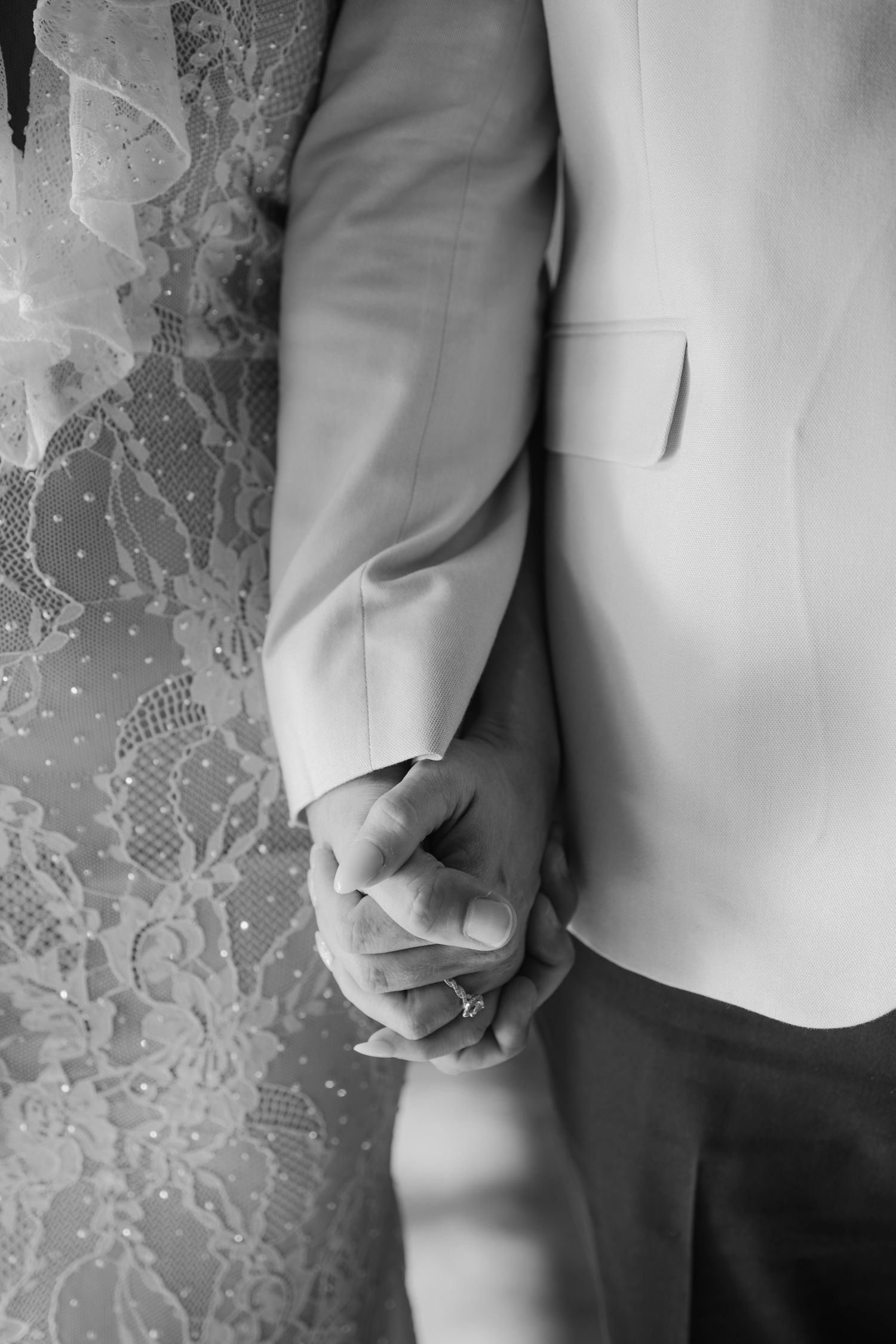 Black and white close-up of bride and groom holding hands, showing wedding ring and lace dress detail.