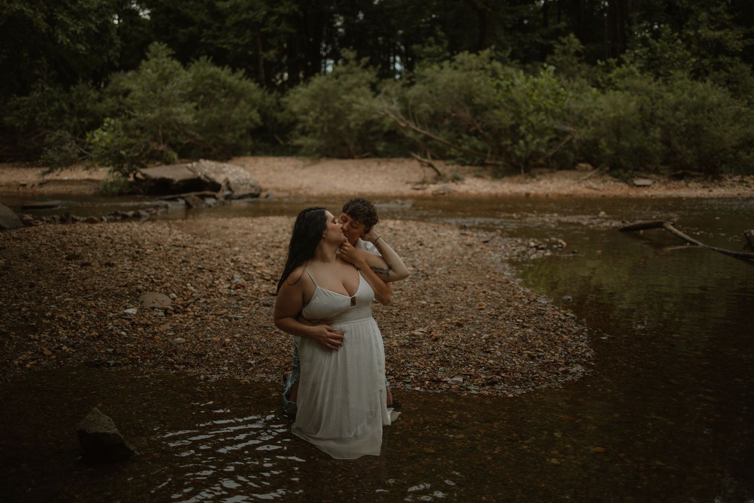 Couple embracing and kissing in shallow river water surrounded by rocky shore and lush green forest trees.
