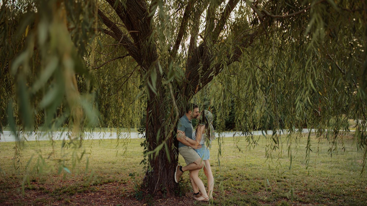 A couple embraces romantically under a weeping willow tree in a lush green park setting.