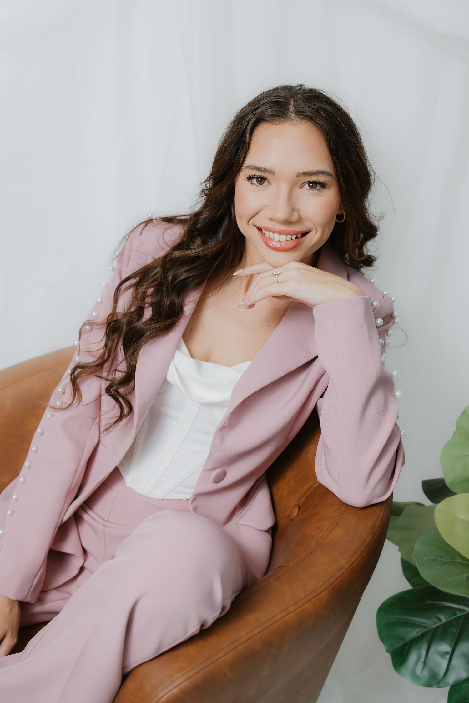 Smiling woman in pink blazer suit seated in leather chair with green plant nearby.