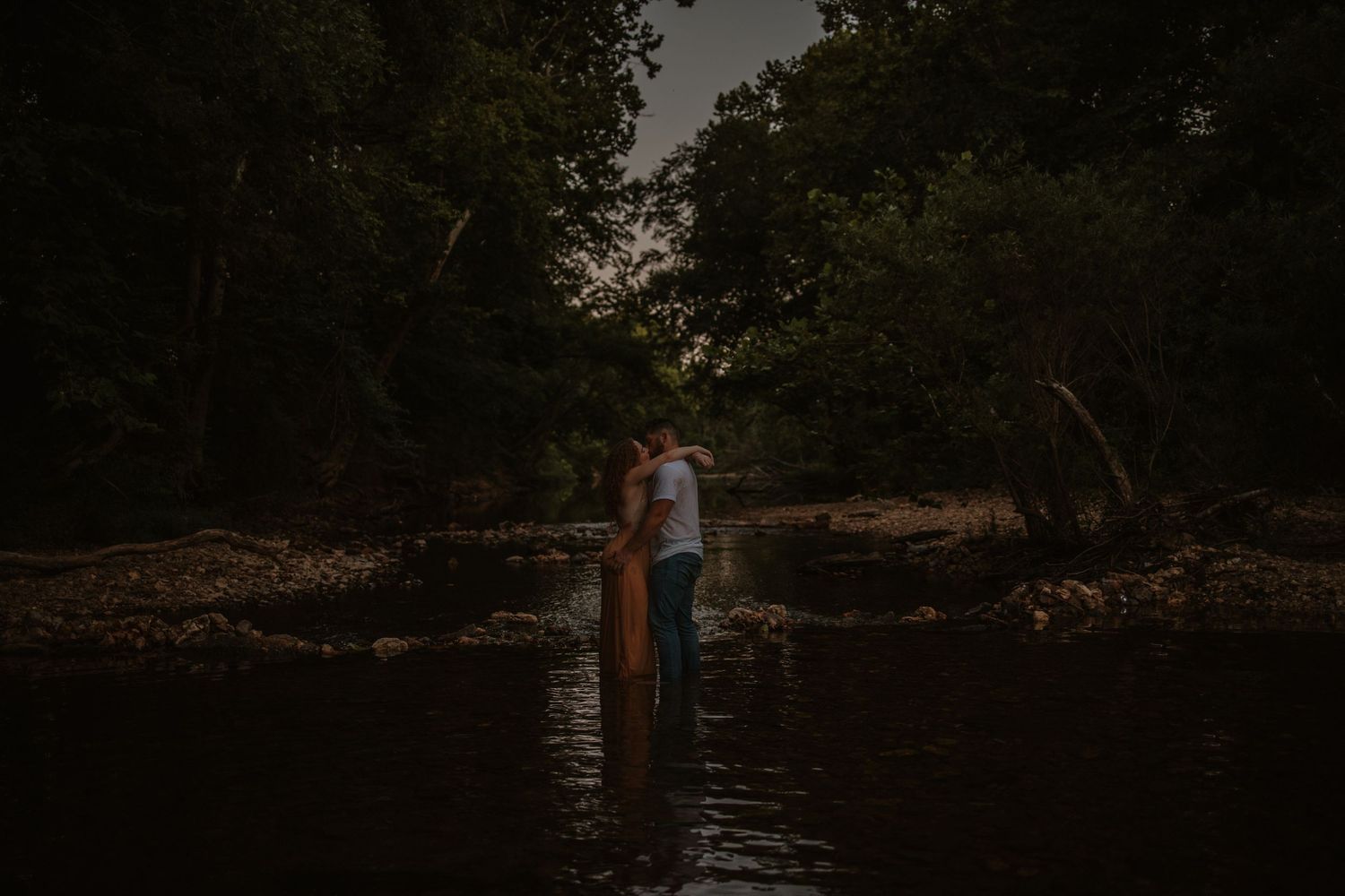 Couple embracing in a dark, moody river surrounded by lush green trees at dusk.