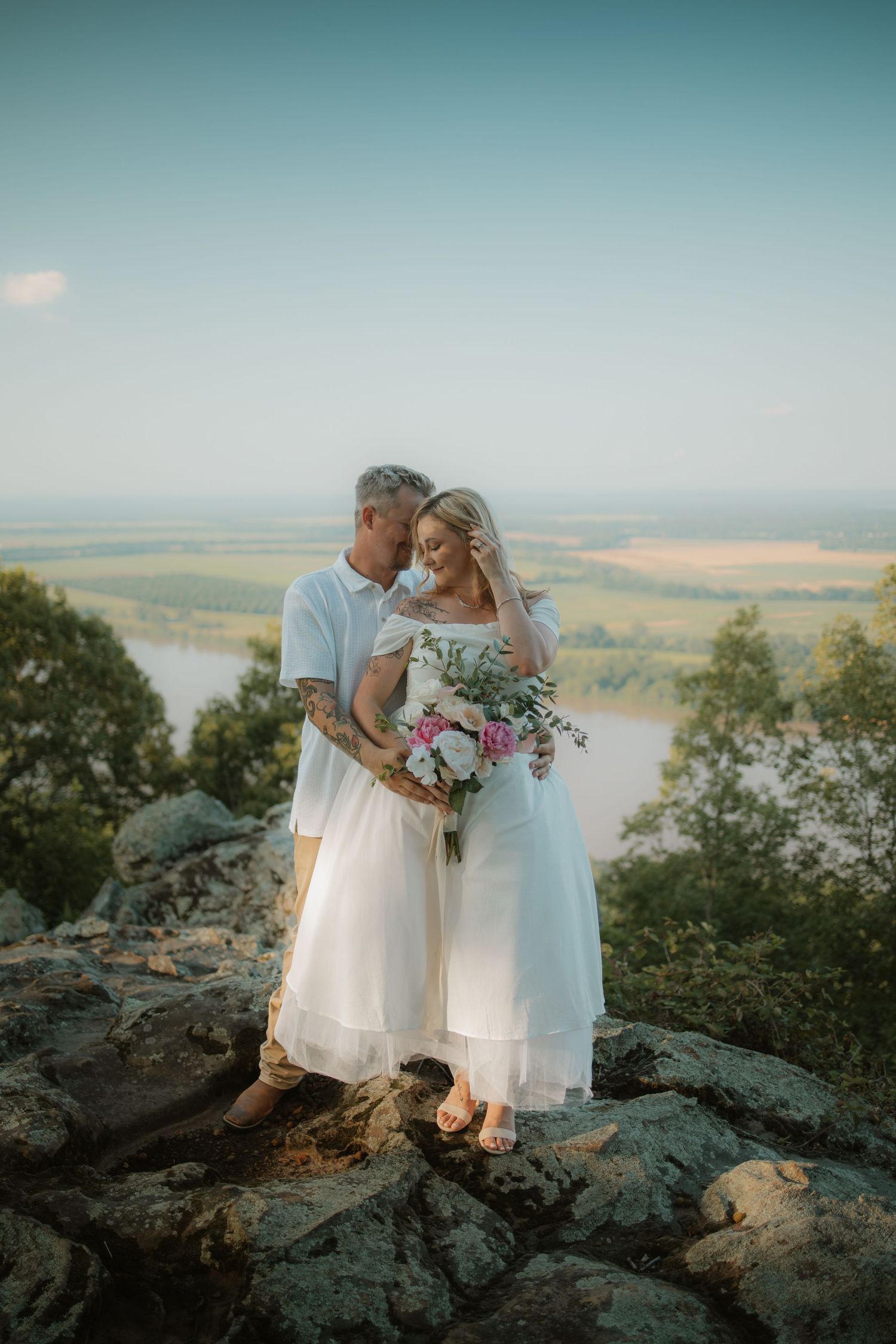 Couple in white wedding attire embracing on rocky overlook with scenic valley view.