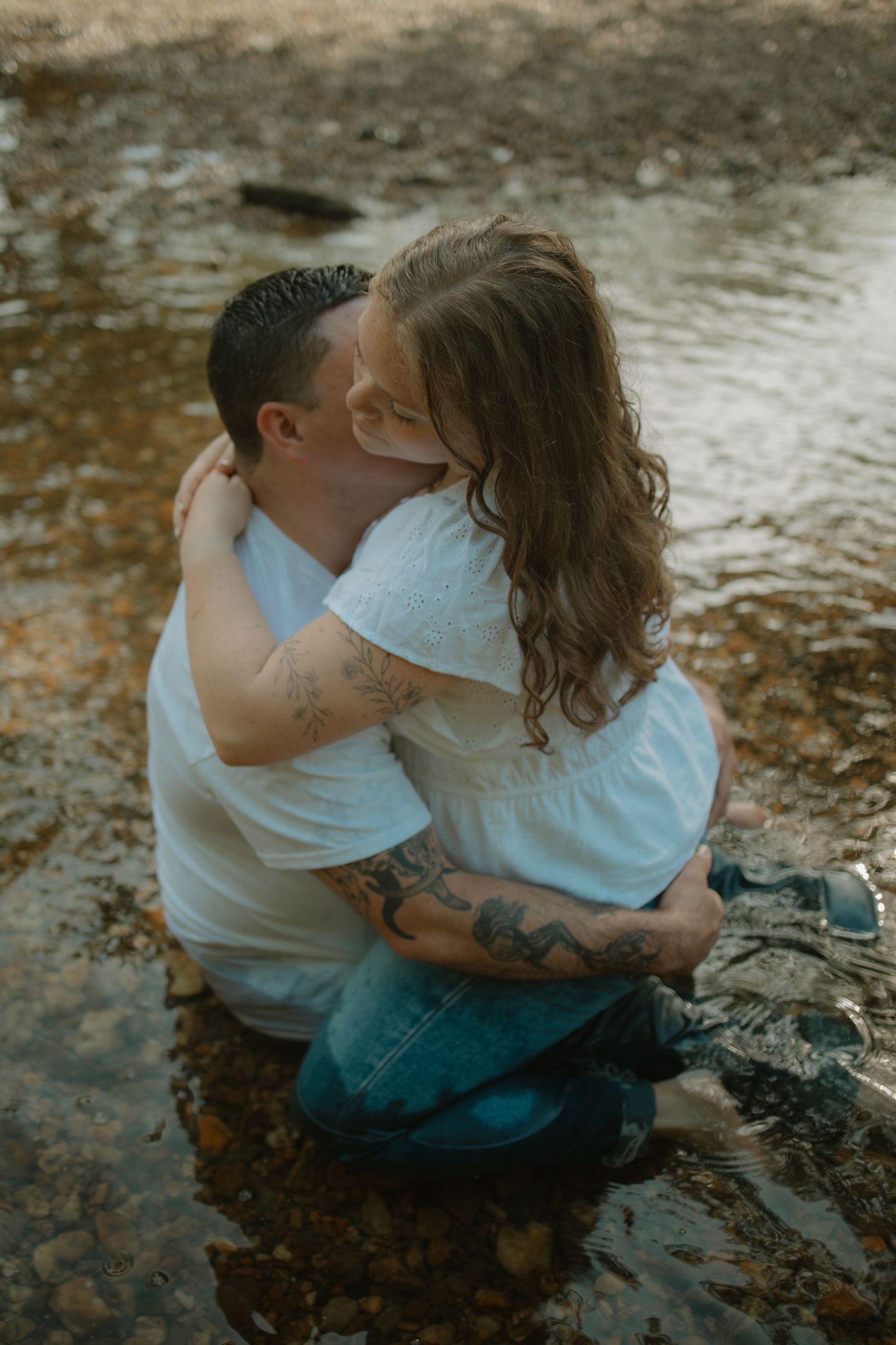 Tattooed couple shares a kiss while crouching in a shallow creek during outdoor photo session.