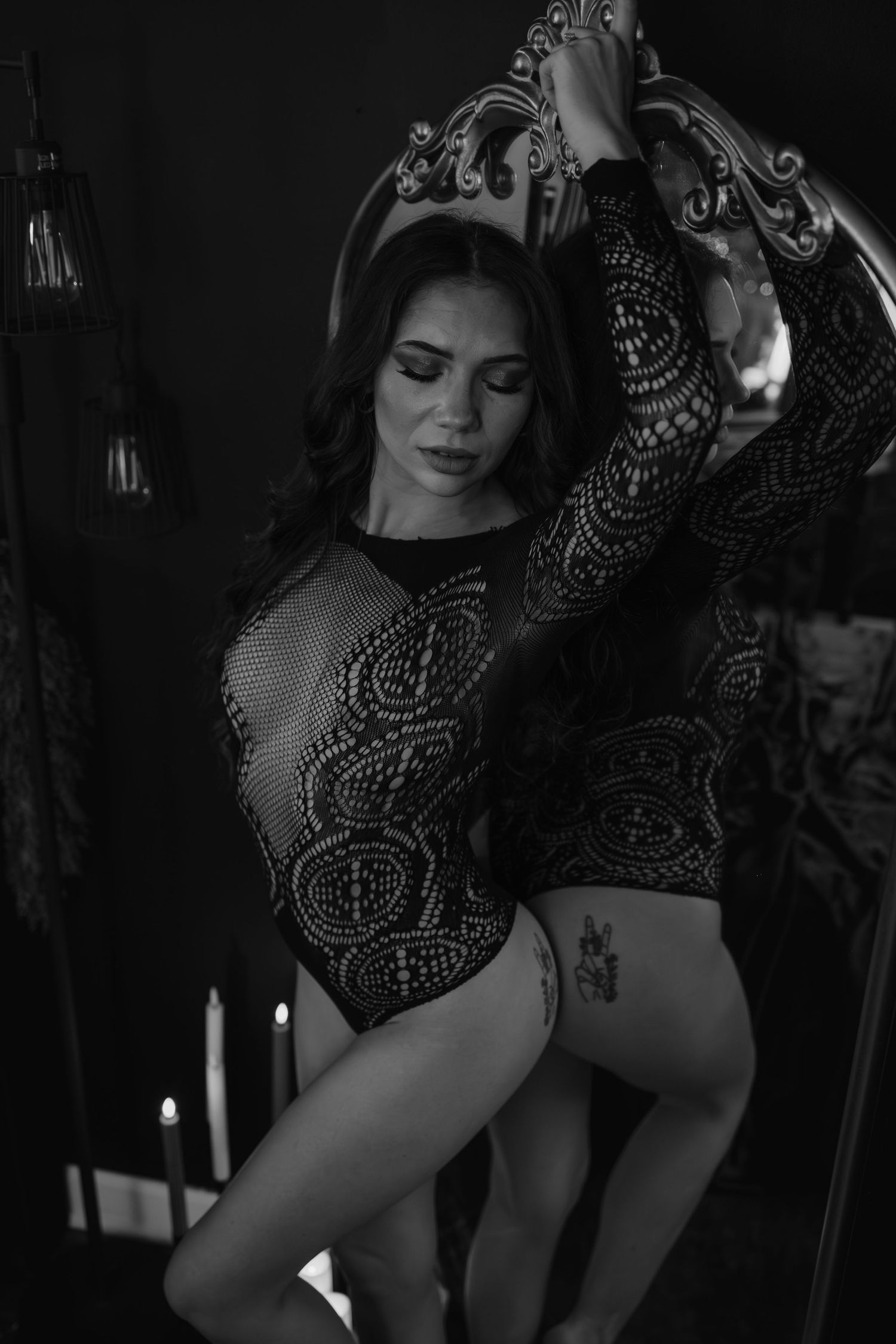 Black and white photo of a woman in lace bodysuit posing elegantly beside an ornate mirror with candles in background.