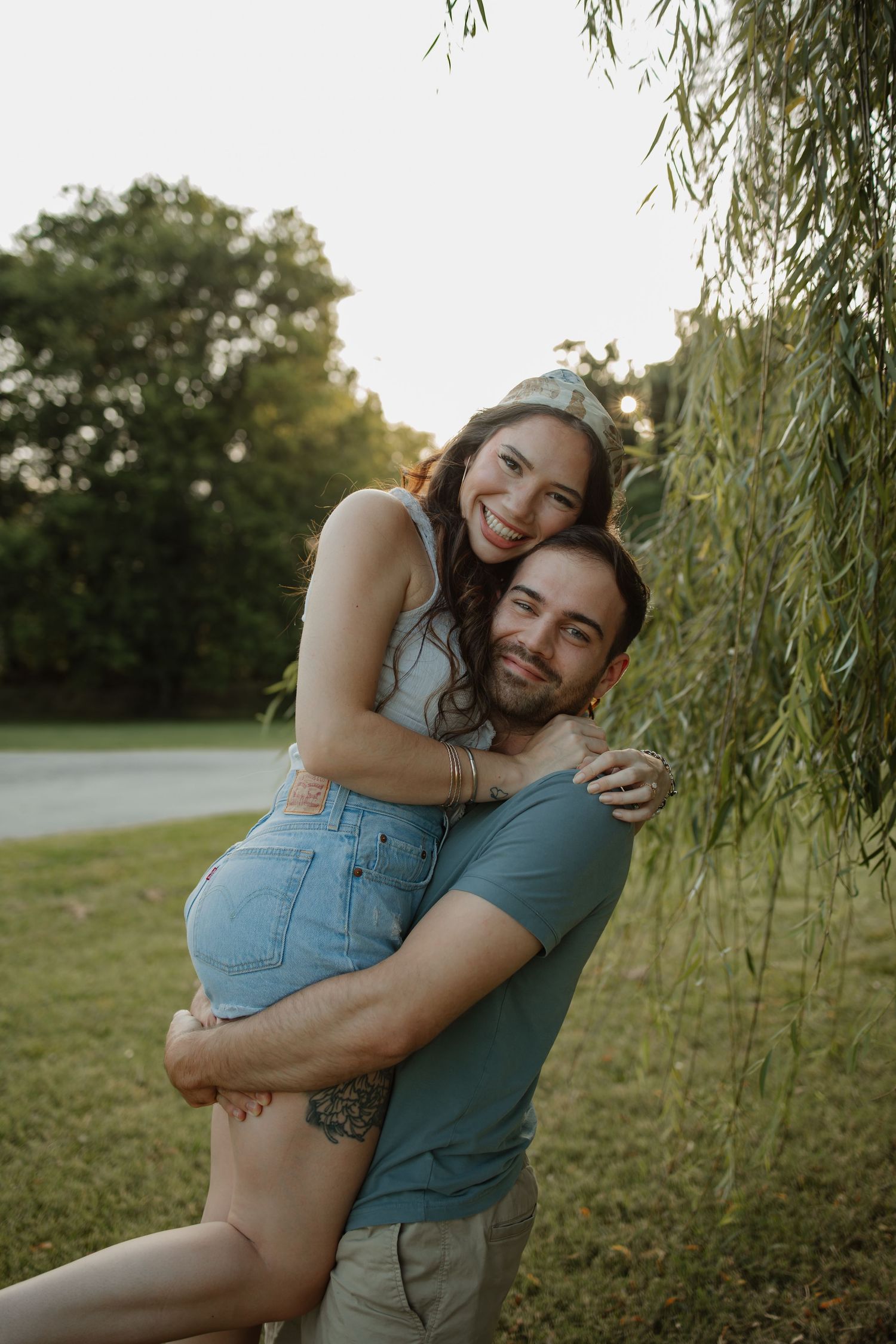 A smiling couple embraces outdoors near a willow tree at sunset, the man lifting the woman in a joyful hug.