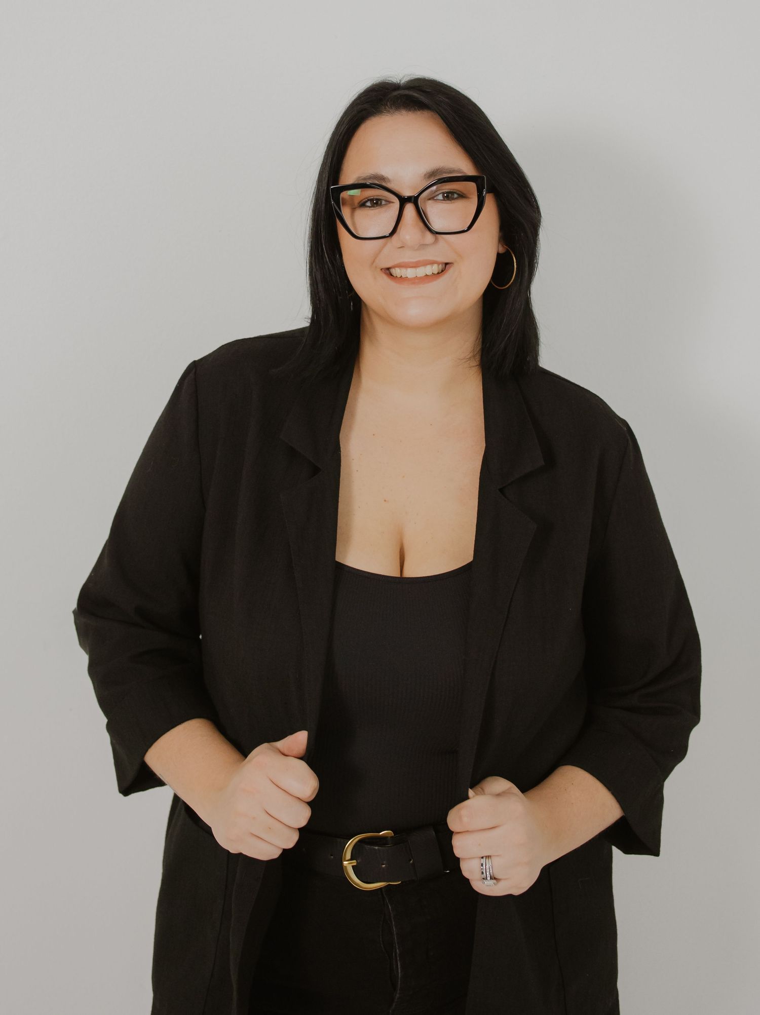 Professional woman in black blazer and glasses smiling confidently against a light gray background.