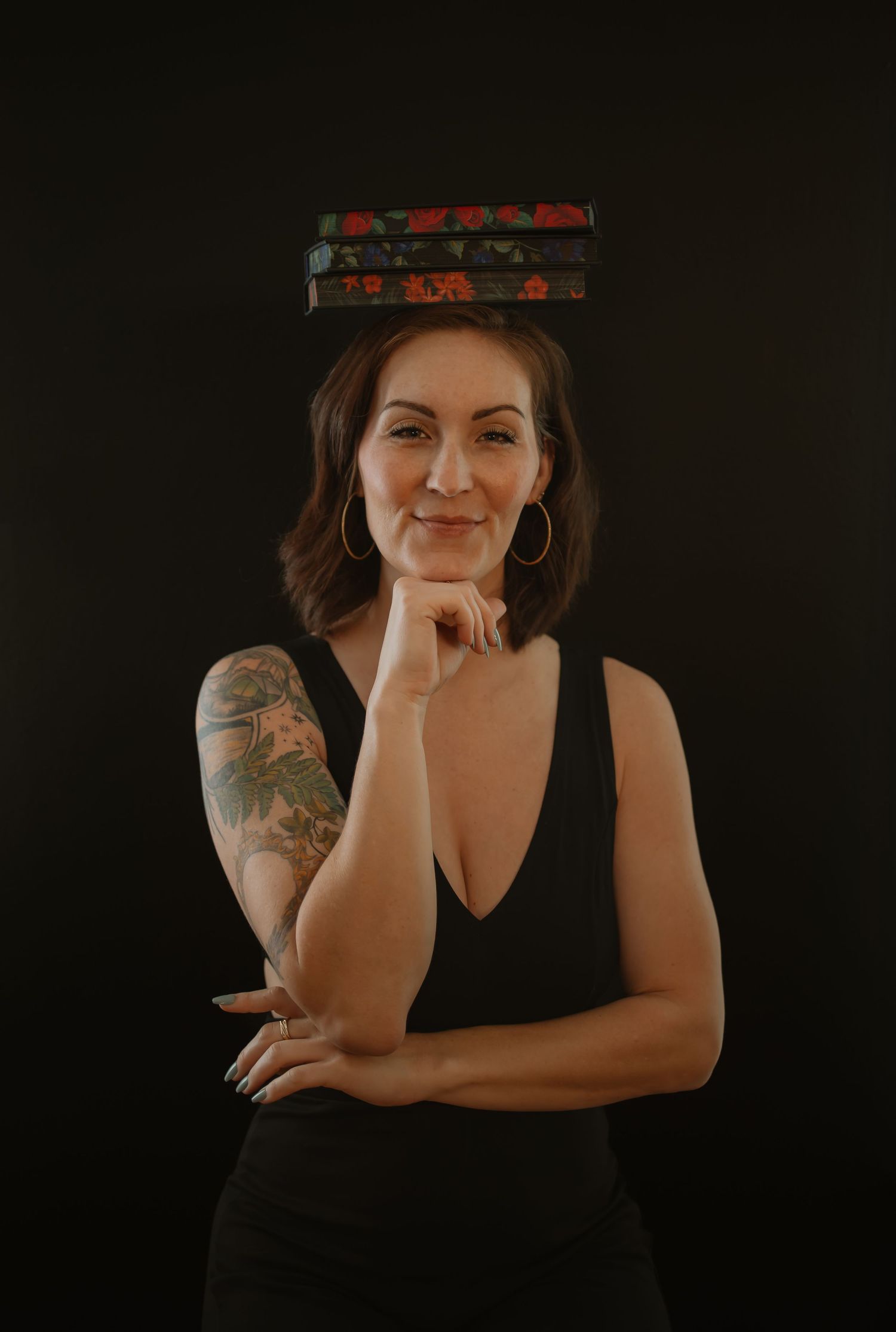 Woman in black dress balancing books on her head, showing arm tattoo against dark background.